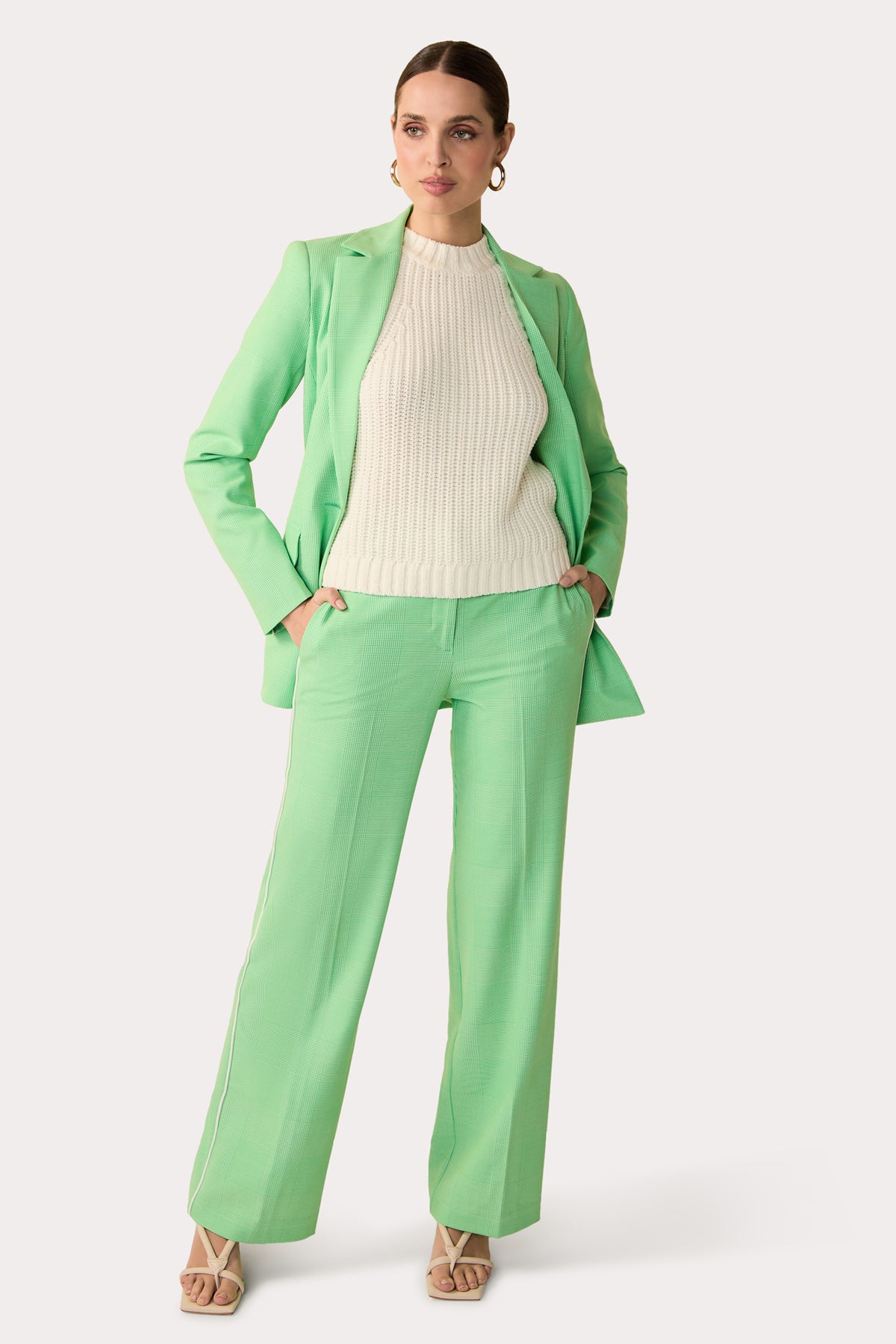 A woman stands confidently in the Bjorn Wide-Leg Pant – Game Time Glen Plaid, paired with a matching blazer over a cream ribbed sweater. She completes her look with beige heeled sandals and hoop earrings, hands in her pockets.