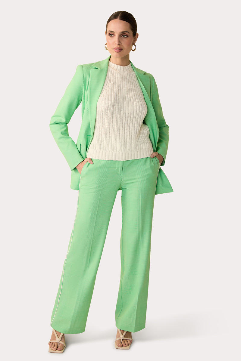A woman stands confidently in the Bjorn Wide-Leg Pant – Game Time Glen Plaid, paired with a matching blazer over a cream ribbed sweater. She completes her look with beige heeled sandals and hoop earrings, hands in her pockets.