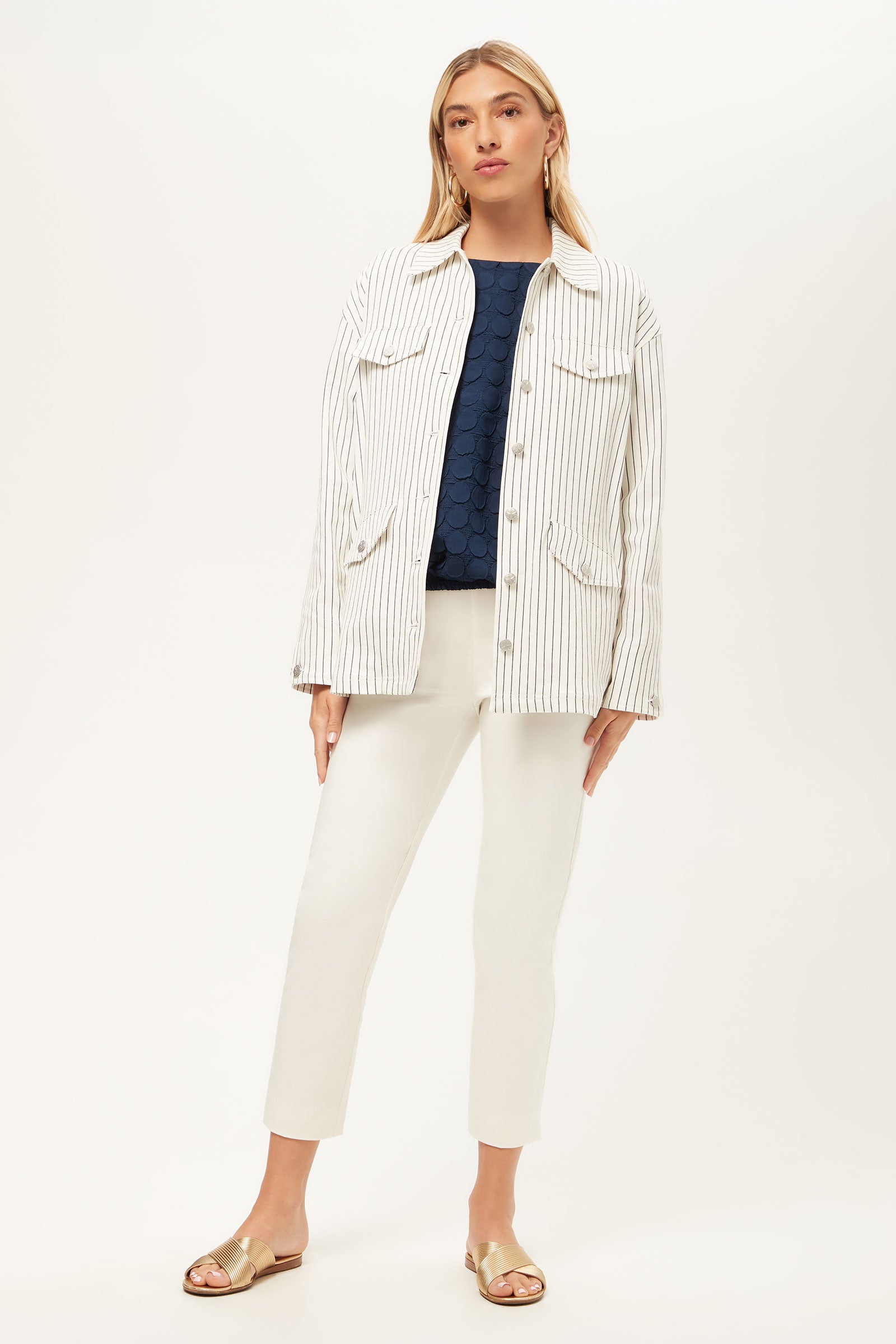 A woman with straight blonde hair poses confidently against a white background, wearing The Game Four-Pocket Jacket in Seles Stripe over a navy top, paired with white pants and gold slide sandals.