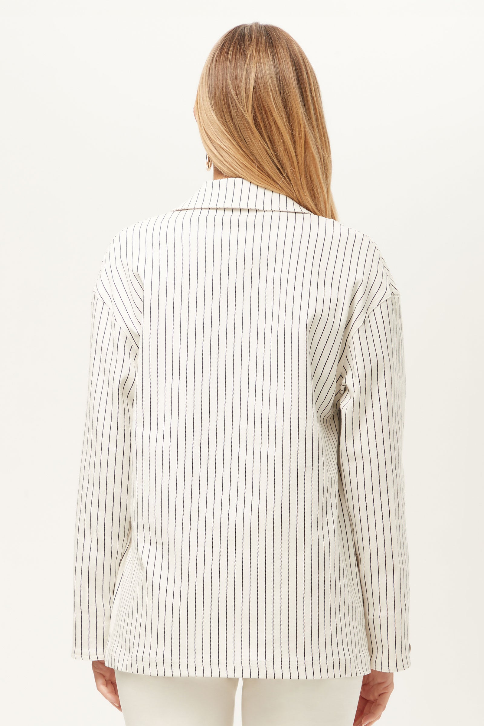 A person with long, light brown hair is seen from behind wearing The Game Four-Pocket Jacket - Seles Stripe, featuring vertical black pinstripes and paired with matching white pants against a plain light background.