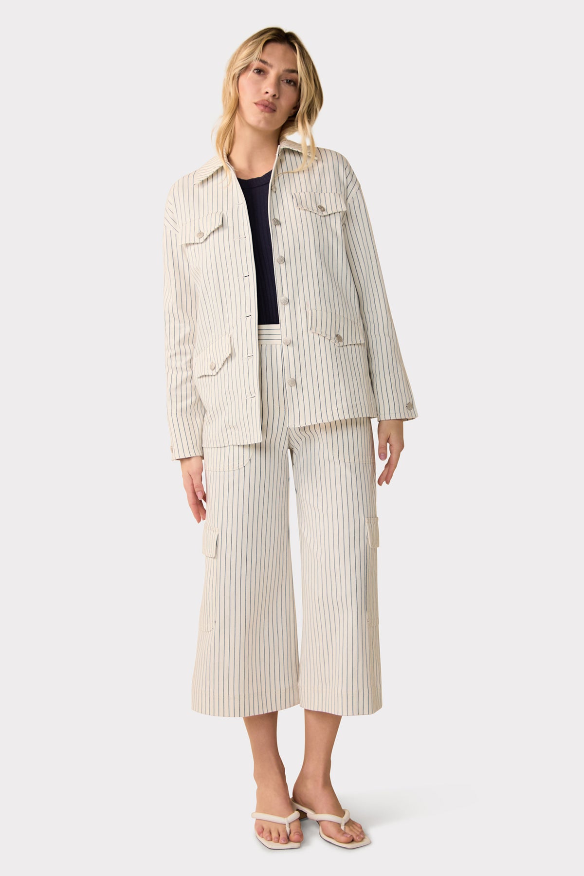 A woman stands in a relaxed pose against a plain light background, wearing the Agassi Wide-Leg Cargo Pant in Seles Stripe with a matching pinstripe jacket, navy top, and white sandals.