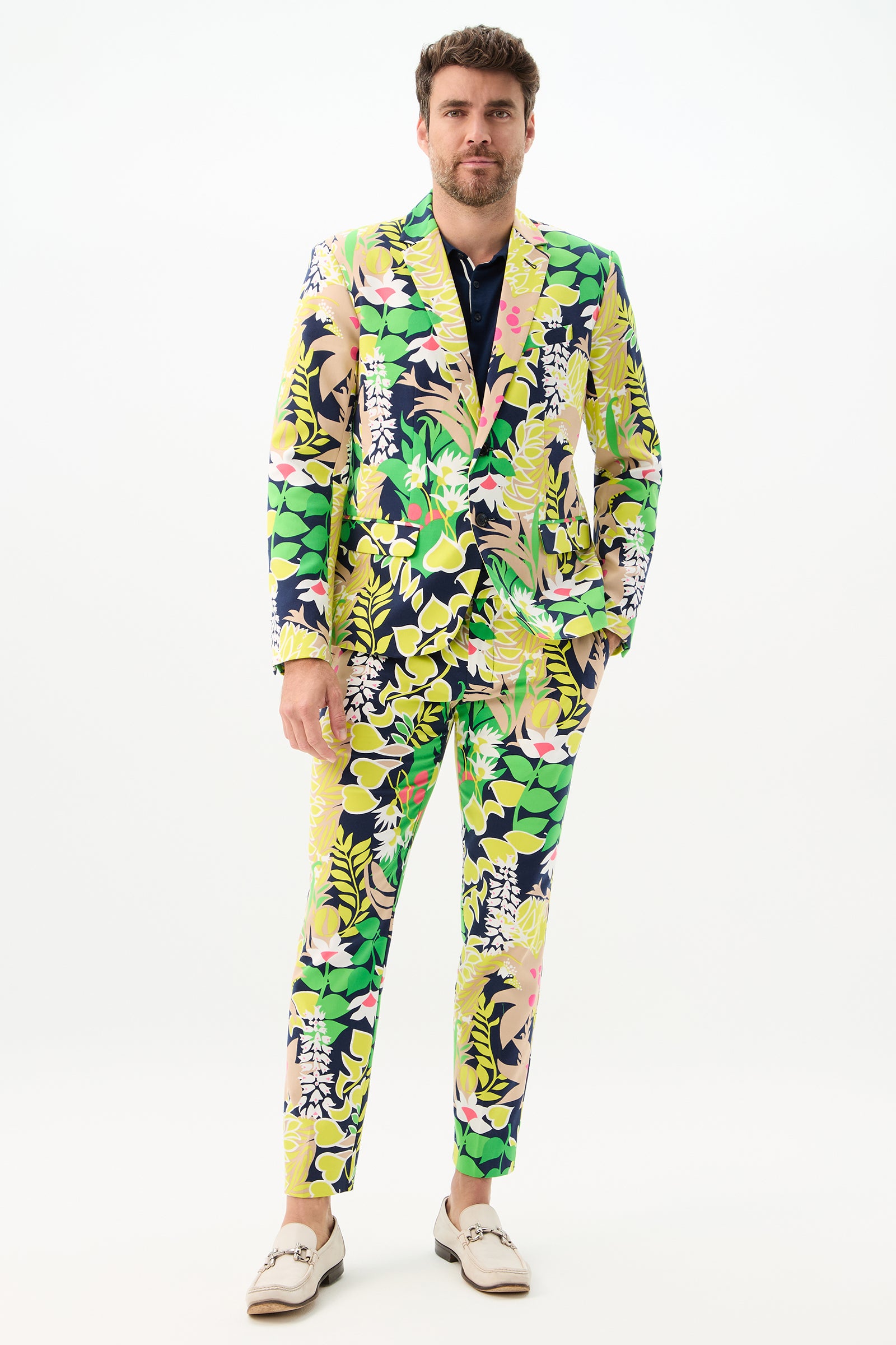 A man faces forward wearing the Thurston Blazer - PS Eternal, featuring a vibrant yellow, green, pink, and navy floral and leaf pattern. He has one hand in his pocket and wears light-colored loafers against a plain white background.