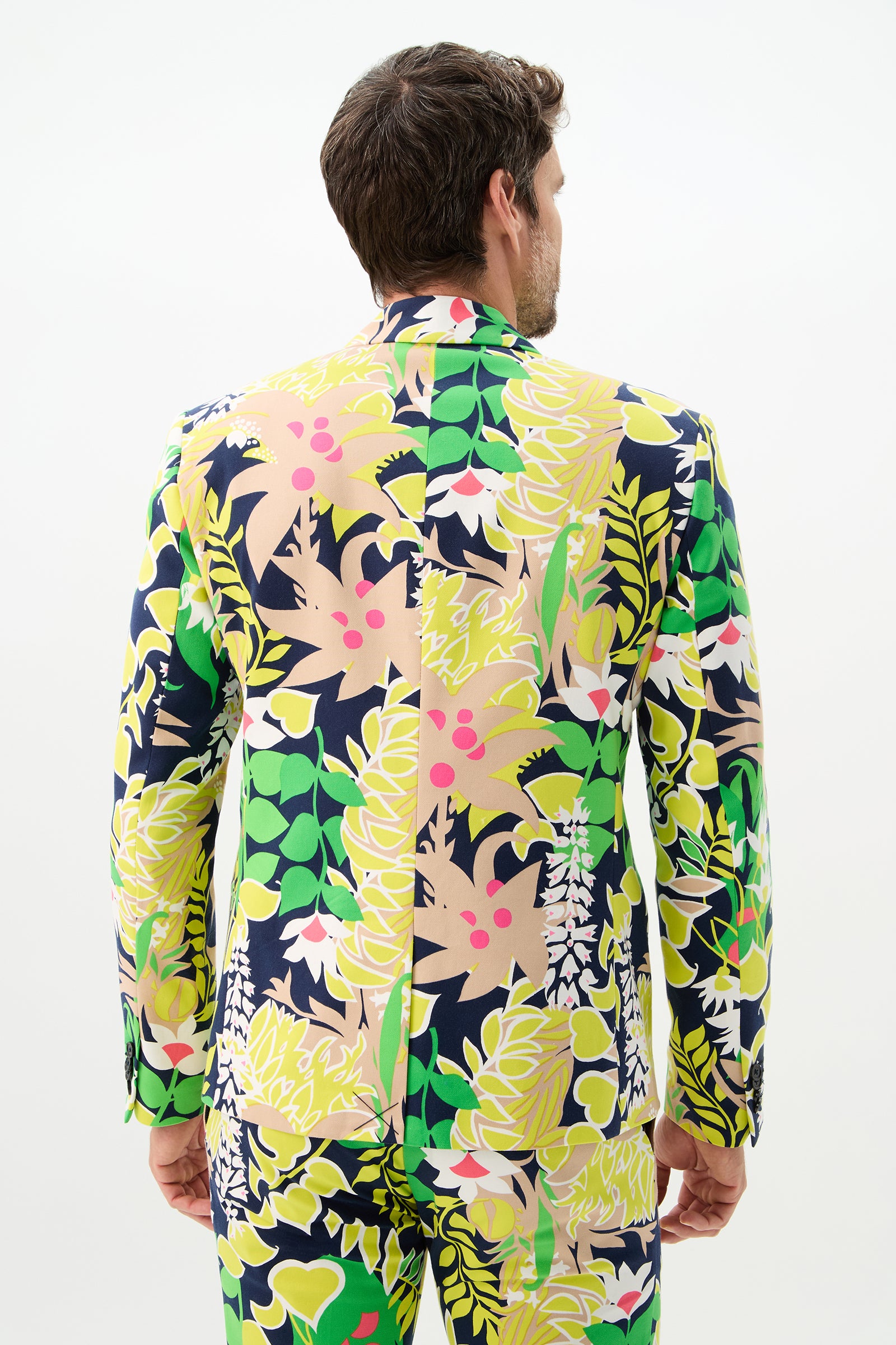 A man faces away, wearing the Thurston Blazer - PS Eternal in Palm Springs Eternal, featuring a bold tropical leaf and floral pattern in green, yellow, pink, beige, and navy against a plain white background.