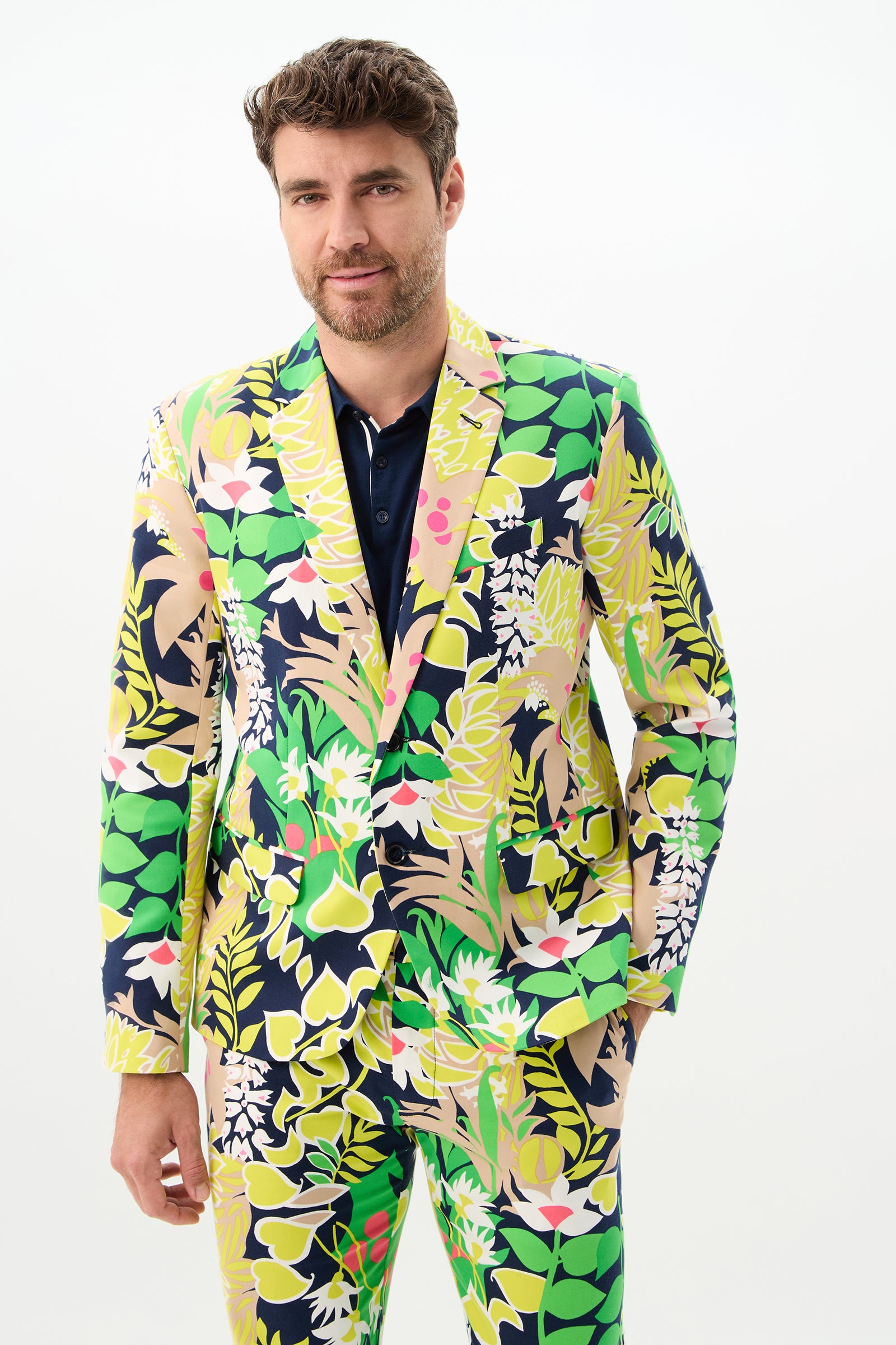 A man with short brown hair and a beard wears the Thurston Blazer - PS Eternal, featuring a bold, tropical floral pattern in green, yellow, pink, and navy. He stands against a plain white background, looking at the camera with a slight smile.
