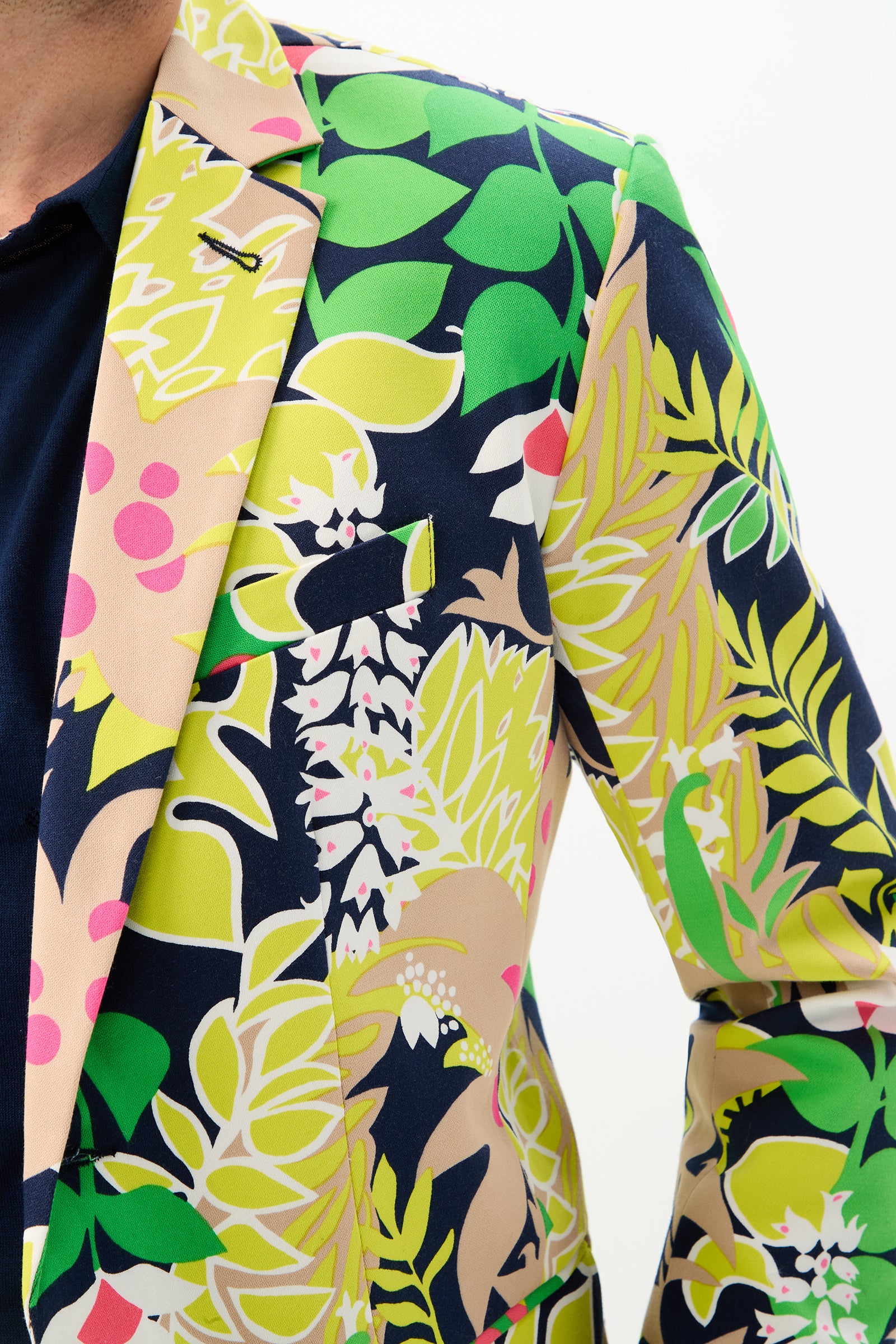A person wears the Thurston Blazer - PS Eternal, featuring a bold tropical floral design in green, yellow, pink, and navy blue. Only their upper body and part of their face are visible against a plain background.