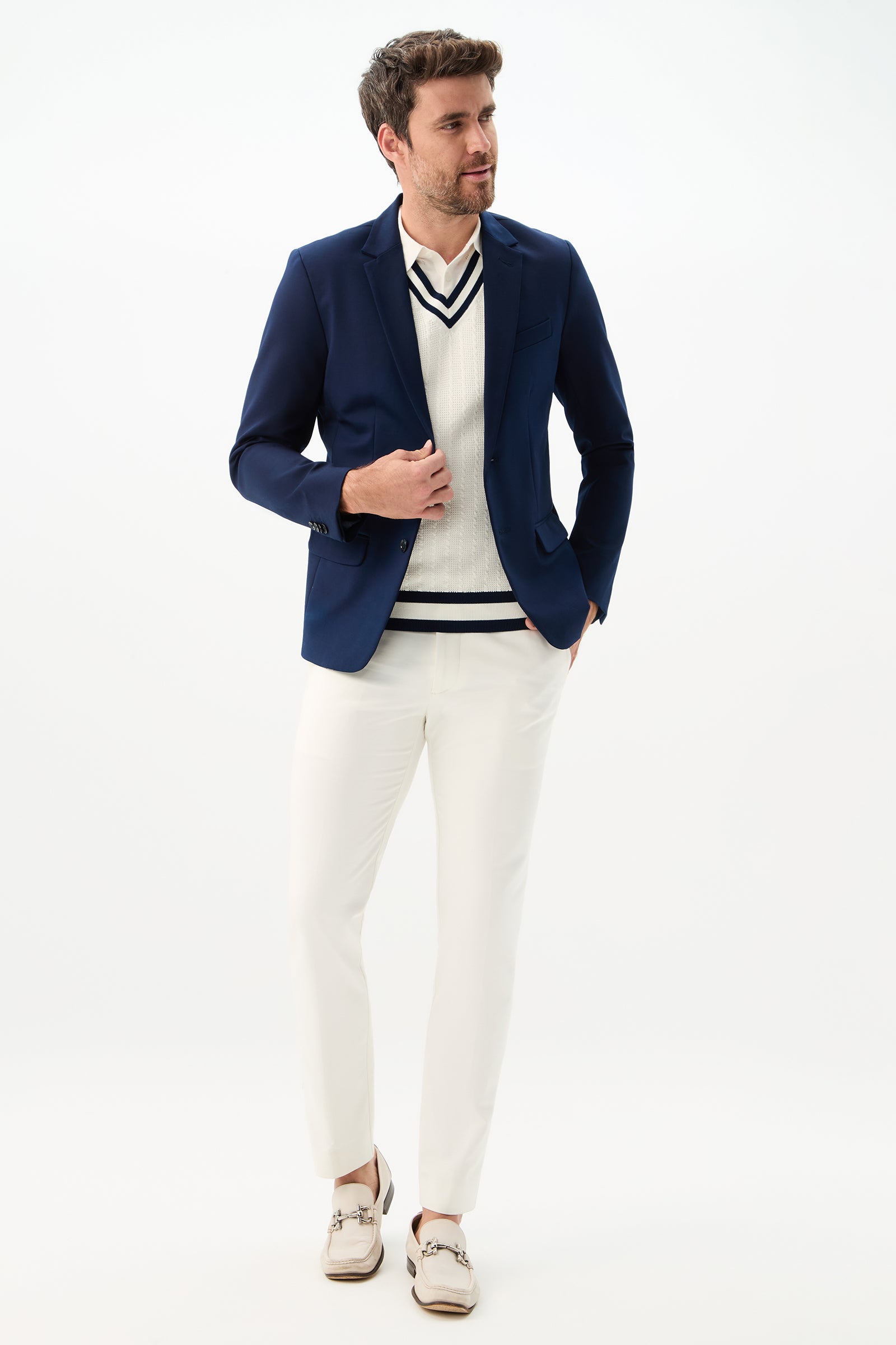 A man stands against a white background, looking to the side as he adjusts his Thurston Blazer - Solid Suiting, worn over a white striped sweater, with white pants and beige loafers.