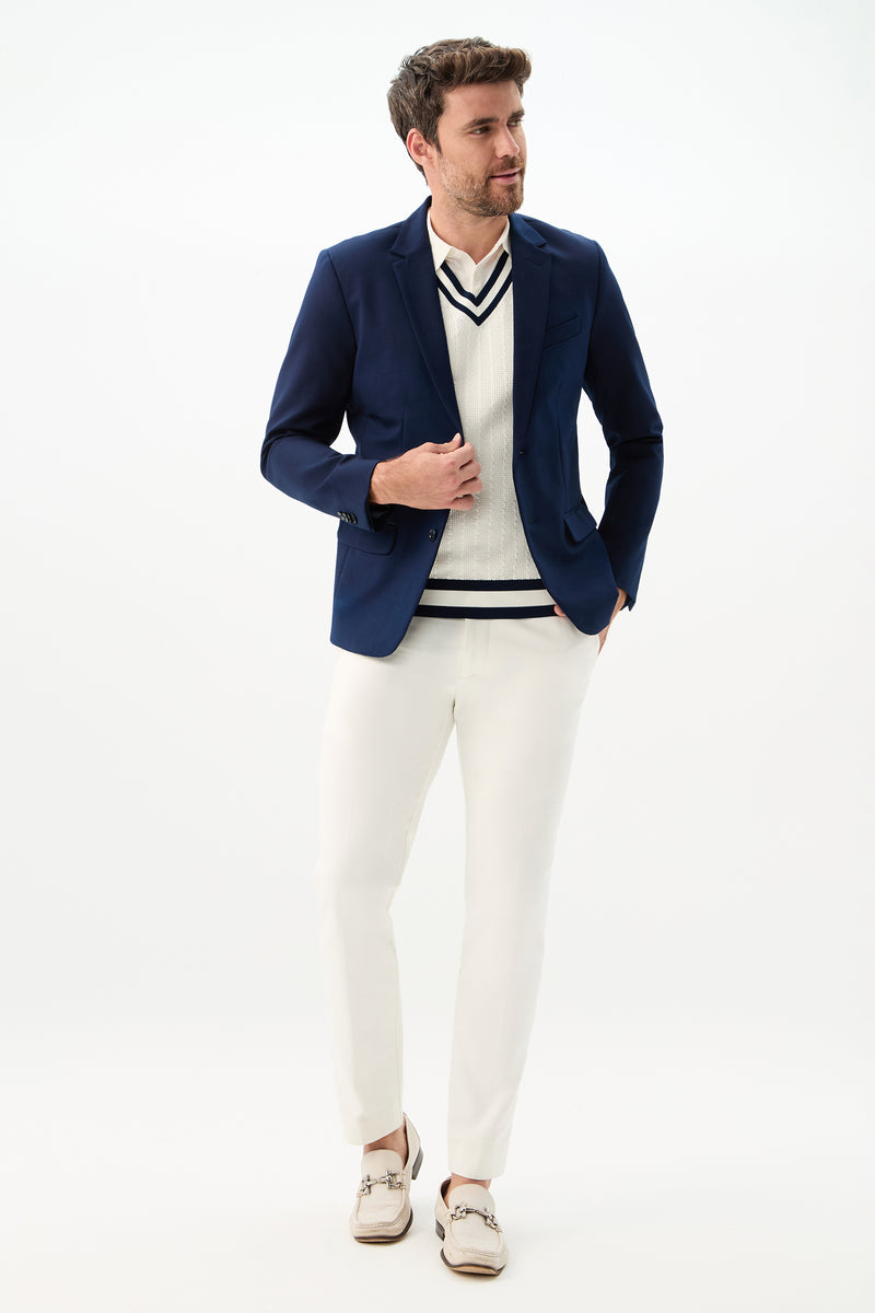 A man wearing white Clyde Slim Trouser - Solid Suiting, a navy blazer, a white V-neck cricket sweater with navy stripes, and beige boat shoes stands against a plain white background.