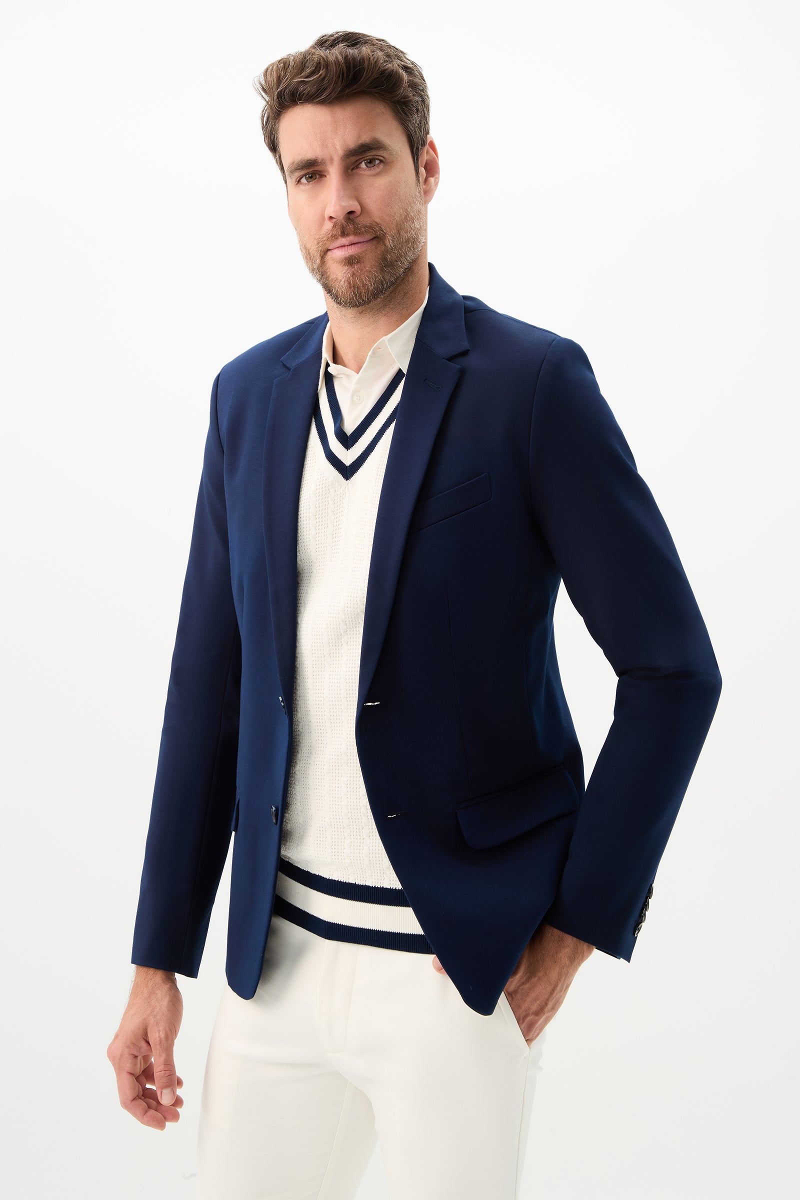 A man with short brown hair and a beard confidently poses in the Thurston Blazer - Solid Suiting, styled over a white sweater with navy trim and white pants, against a plain white background.