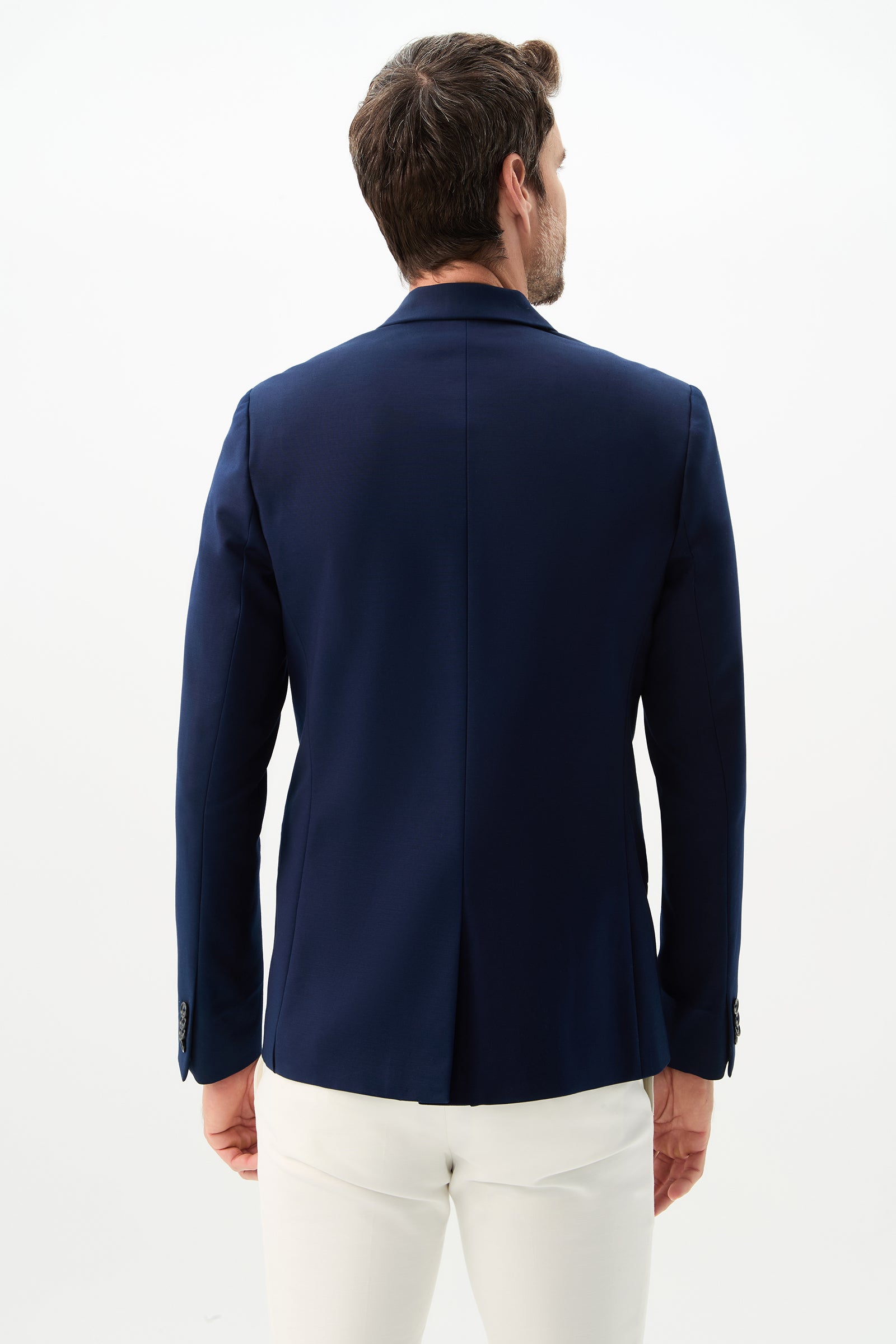 A man is seen from behind wearing the Thurston Blazer - Solid Suiting in navy blue with white pants, standing against a plain white background.