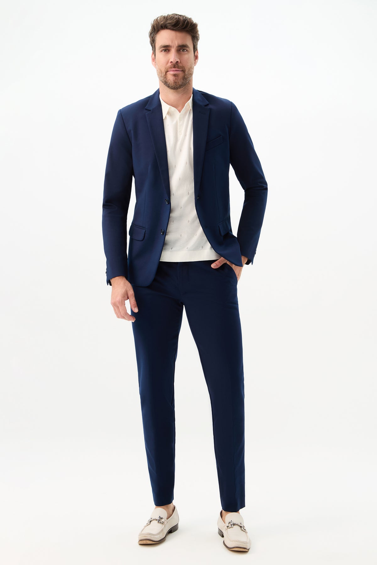 A man with short brown hair and a beard stands against a light background wearing the Thurston Blazer - Solid Suiting over a white shirt with beige loafers. His left hand rests in his pocket.