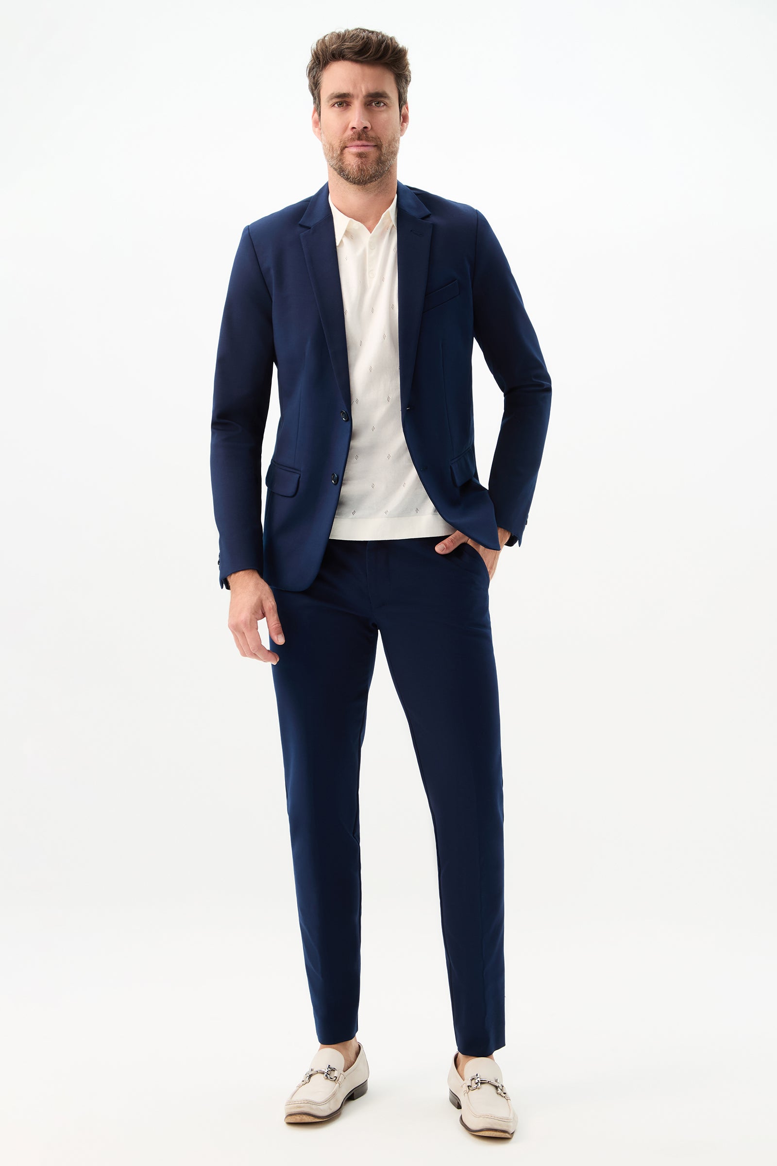 A man with short brown hair and a beard stands against a light background wearing the Thurston Blazer - Solid Suiting over a white shirt with beige loafers. His left hand rests in his pocket.