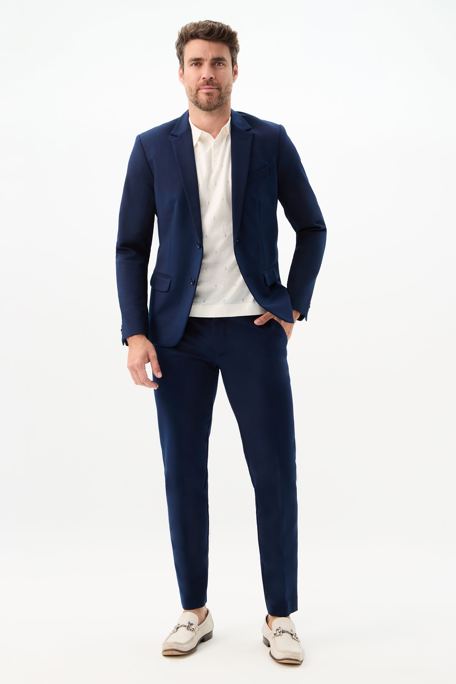 A man with short brown hair and a trimmed beard wears the Clyde Slim Trouser - Solid Suiting in navy, paired with a white shirt and light loafers. He poses against a white background, hand in pocket, with a neutral expression.
