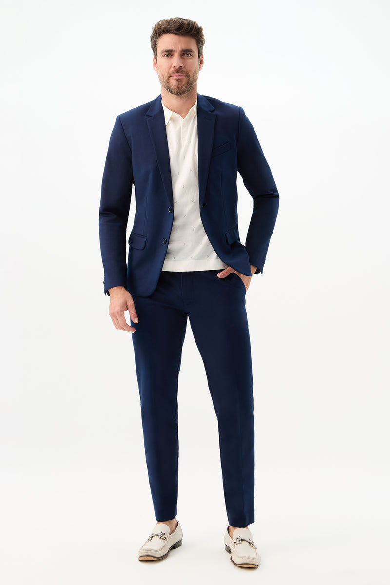 A man with short brown hair and a trimmed beard wears the Clyde Slim Trouser - Solid Suiting in navy, paired with a white shirt and light loafers. He poses against a white background, hand in pocket, with a neutral expression.