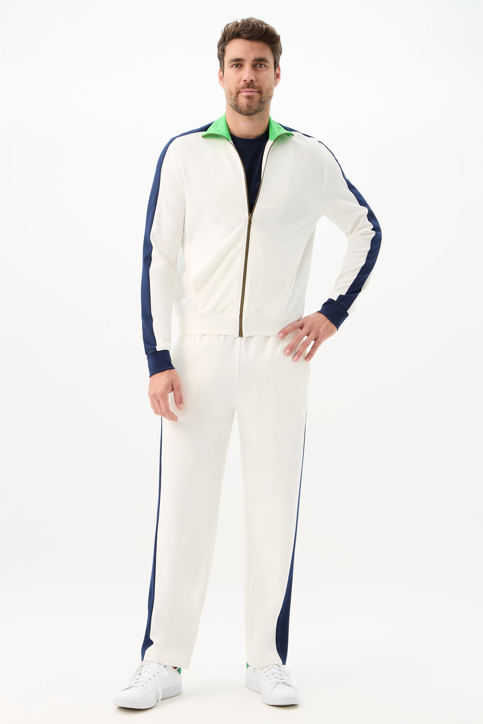 A man faces forward, wearing the Ugo Track Jacket - Cozy Ponte in white with blue side stripes and a green collar over a dark shirt, paired with white sneakers. The background is plain white.