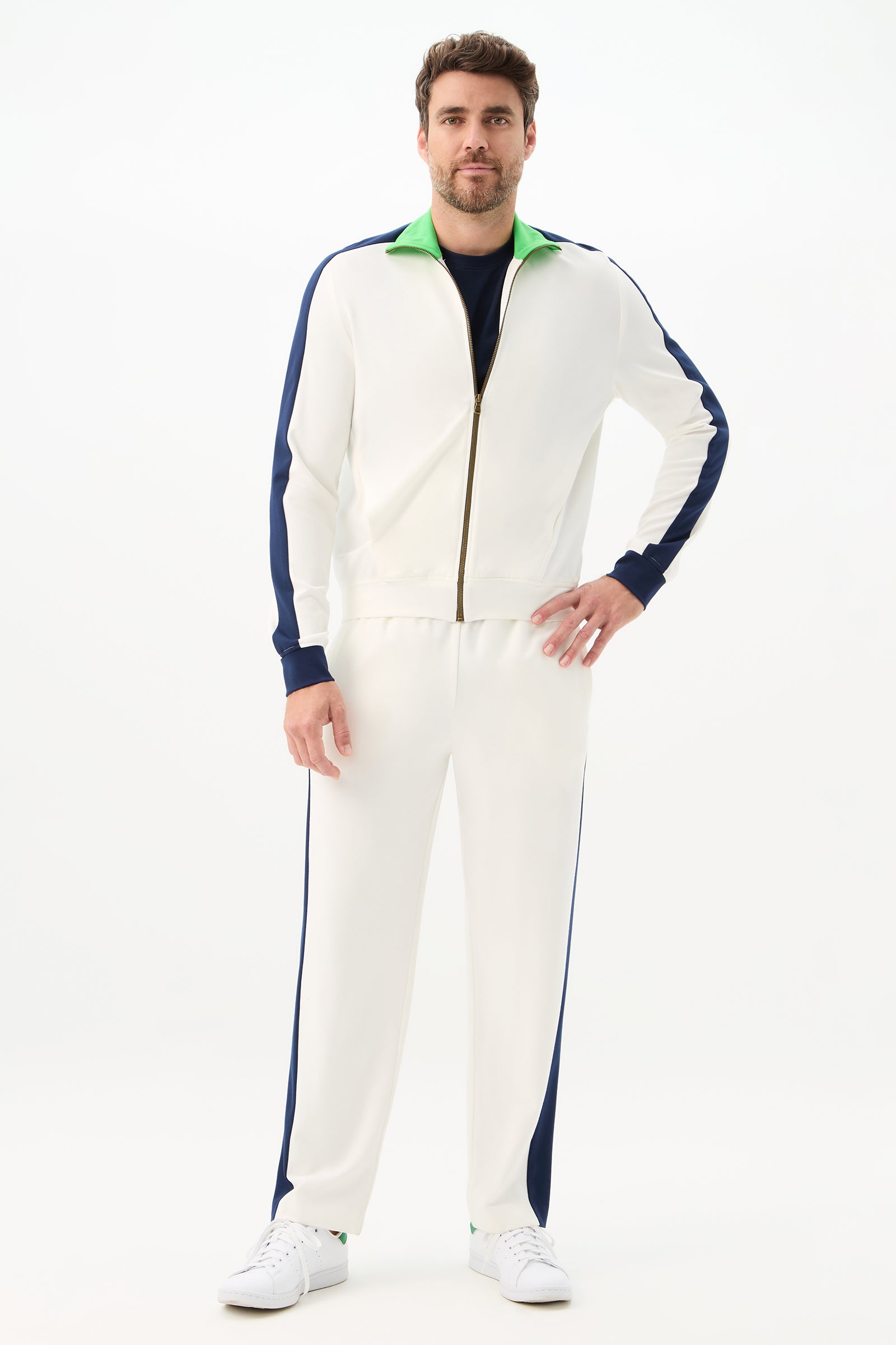 A man facing forward wears the Tennis Track Pant - Cozy Ponte, a white track suit with blue side stripes and a green collar, paired with white sneakers against a plain white background.