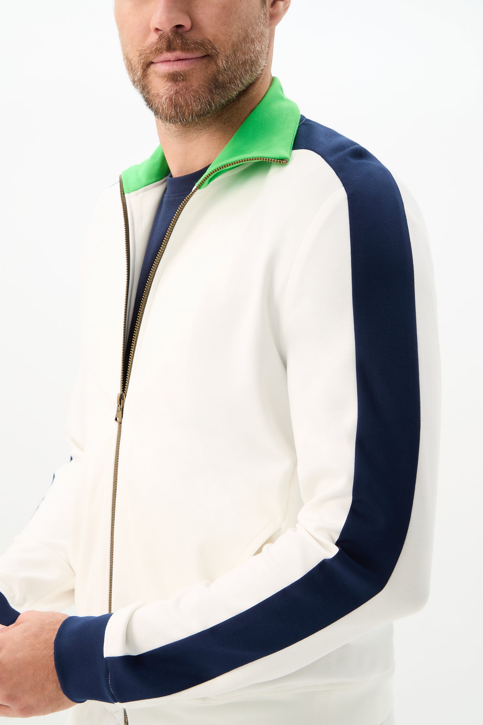 A man models the Ugo Track Jacket - Cozy Ponte, featuring a white zip-up front, navy blue sleeves, and a green collar. He stands against a plain white background with only his lower face and upper body visible.