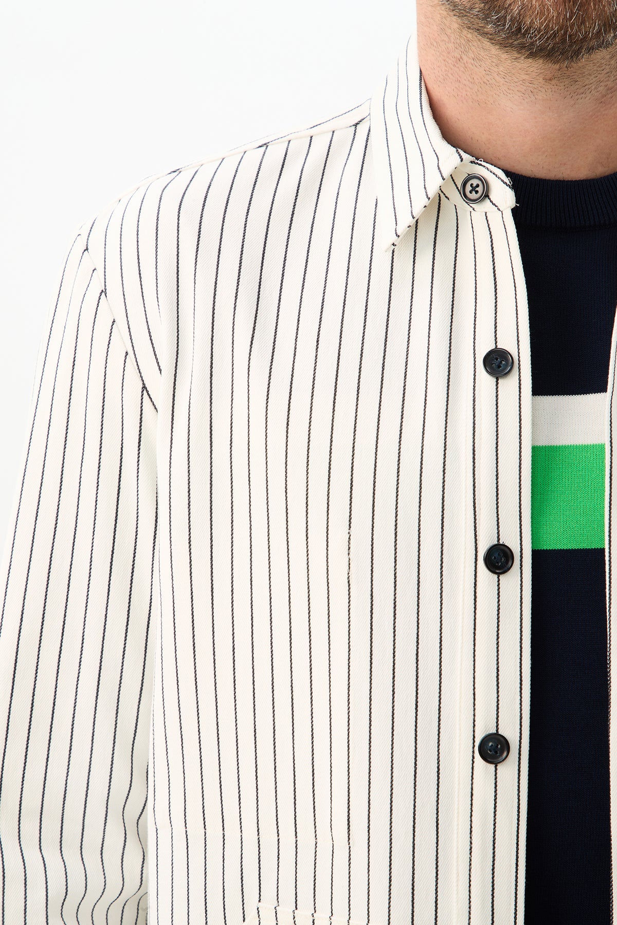A close-up of a person wearing the Mulholland Shirt Jacket - Seles Stripe, a white shirt with black vertical stripes, over a dark sweater with white and green horizontal stripes. The shirt is unbuttoned at the top, partially revealing a bearded face.