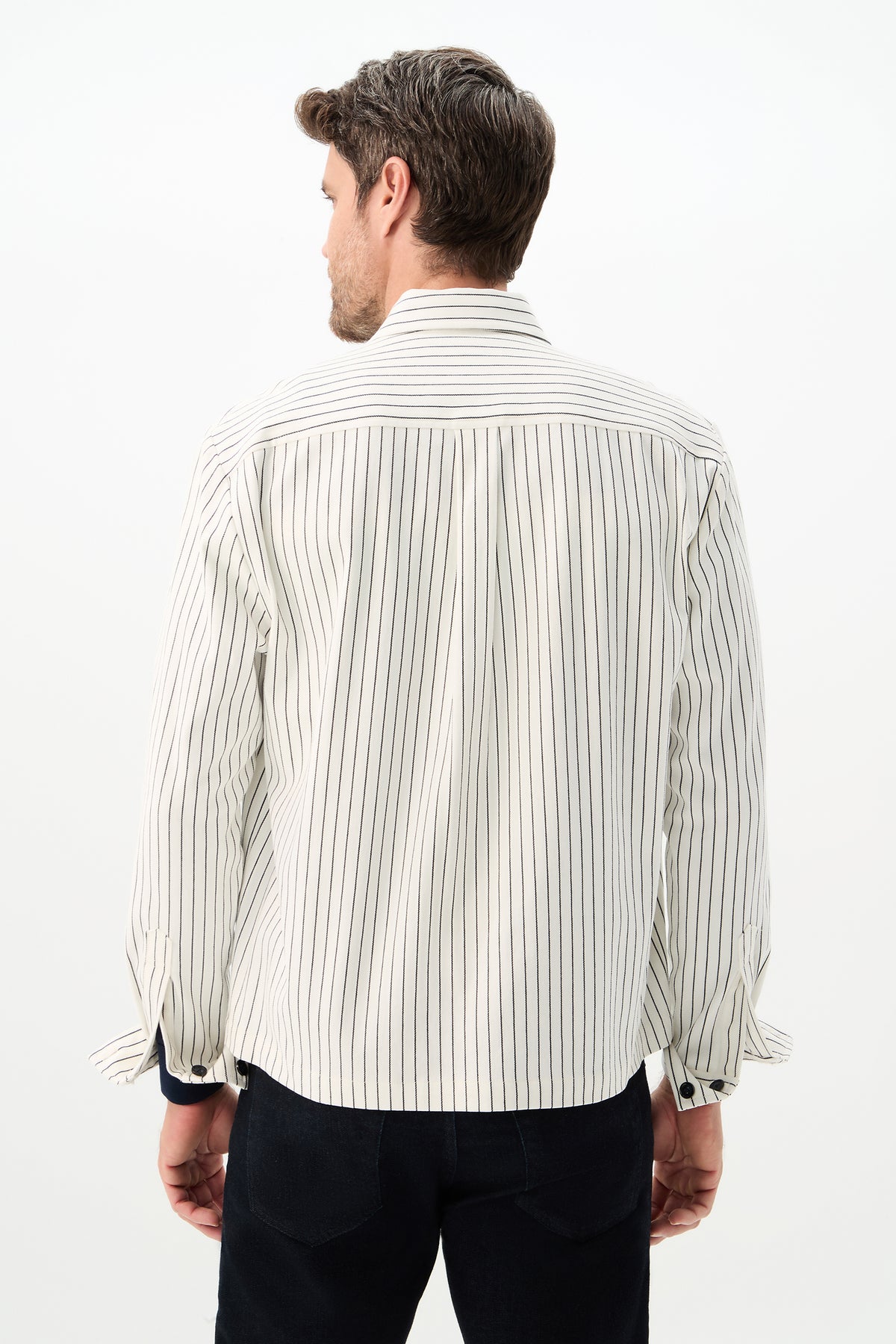 A man faces away from the camera, wearing the Mulholland Shirt Jacket in Seles Stripe—a white shirt jacket with thin black vertical stripes—paired with dark pants, set against a plain white background.