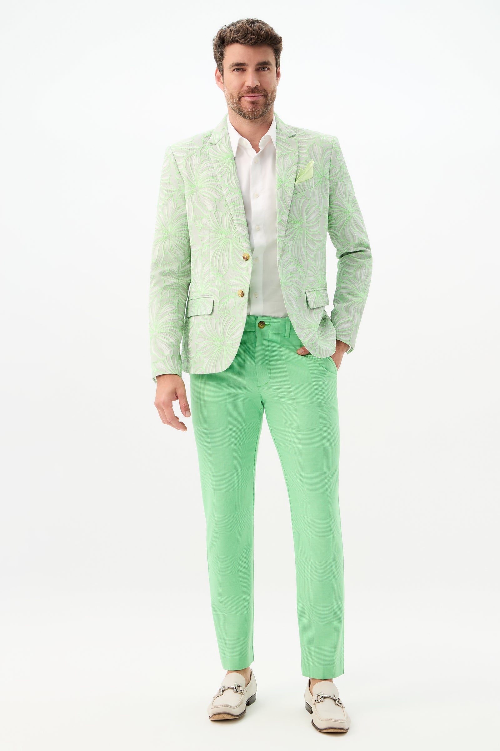 A man with dark hair and a short beard stands against a white background wearing the Thurston Blazer - Hibiscus Jacquard, paired with matching light green trousers, a white shirt, and white loafers.