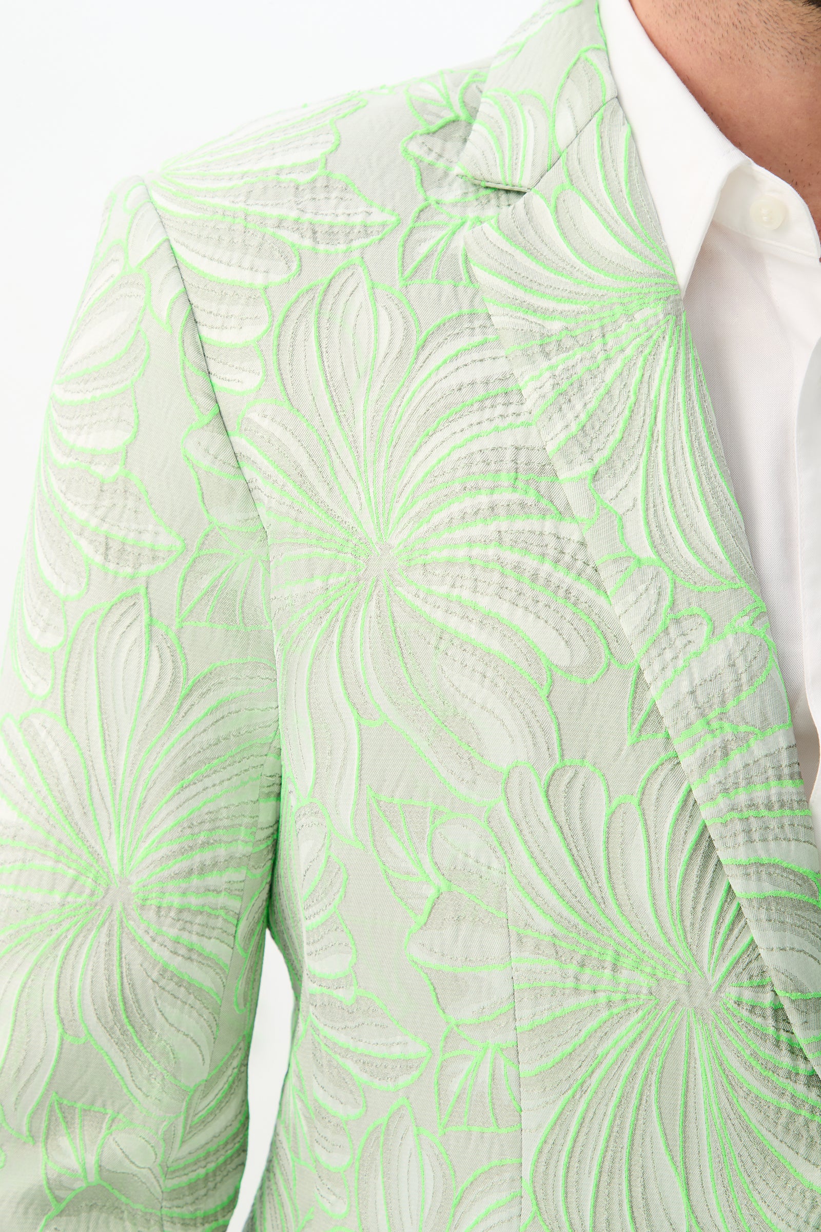 Close-up of a person wearing the Thurston Blazer - Hibiscus Jacquard, featuring an intricate white and neon green floral pattern over light green fabric, partially styled with a white collared shirt. Only the shoulder and part of the chest are visible.