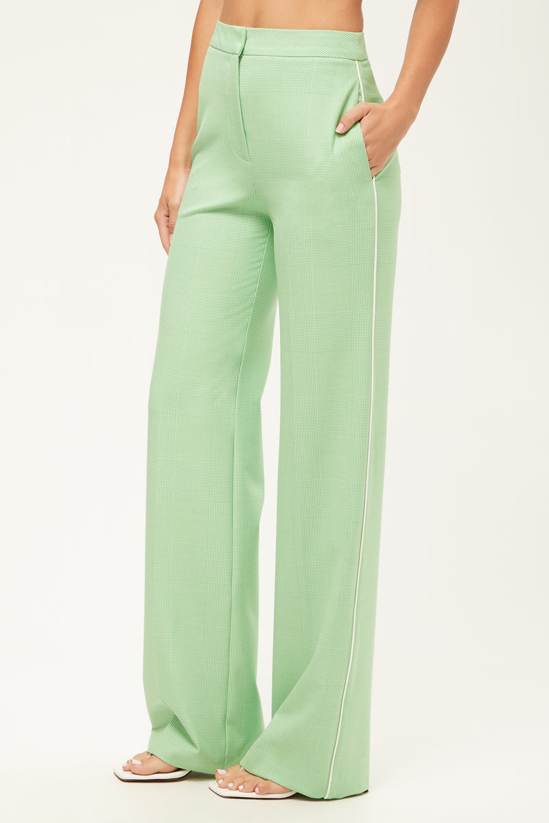 A woman models the Bjorn Wide-Leg Pant in Grass Court Green/Whitewash with white piping, hands in pockets, paired with white sandals against a plain background.