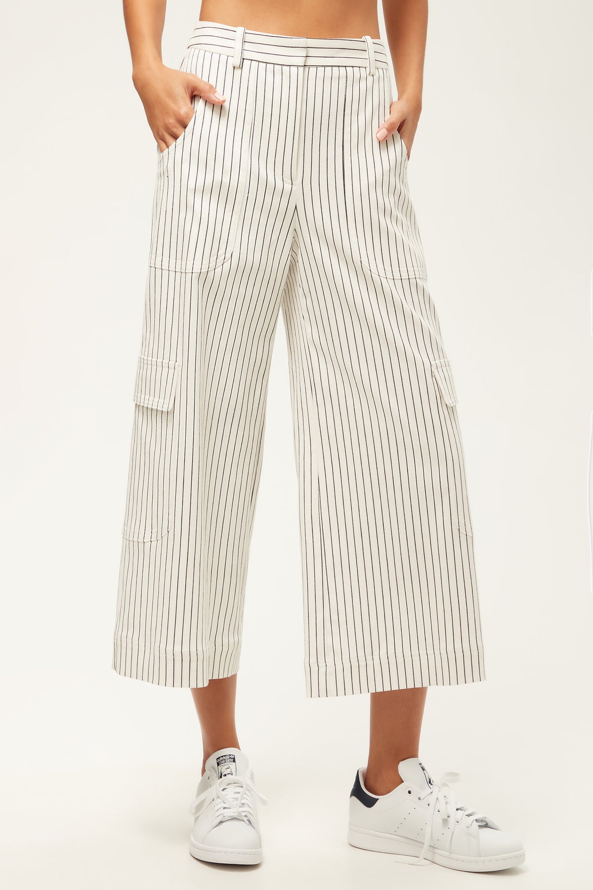 A person wears the Agassi Wide-Leg Cargo Pant - Seles Stripe in Whitewash/Ink—high-waisted, relaxed fit, cropped with black vertical stripes and side pockets—paired with white sneakers.