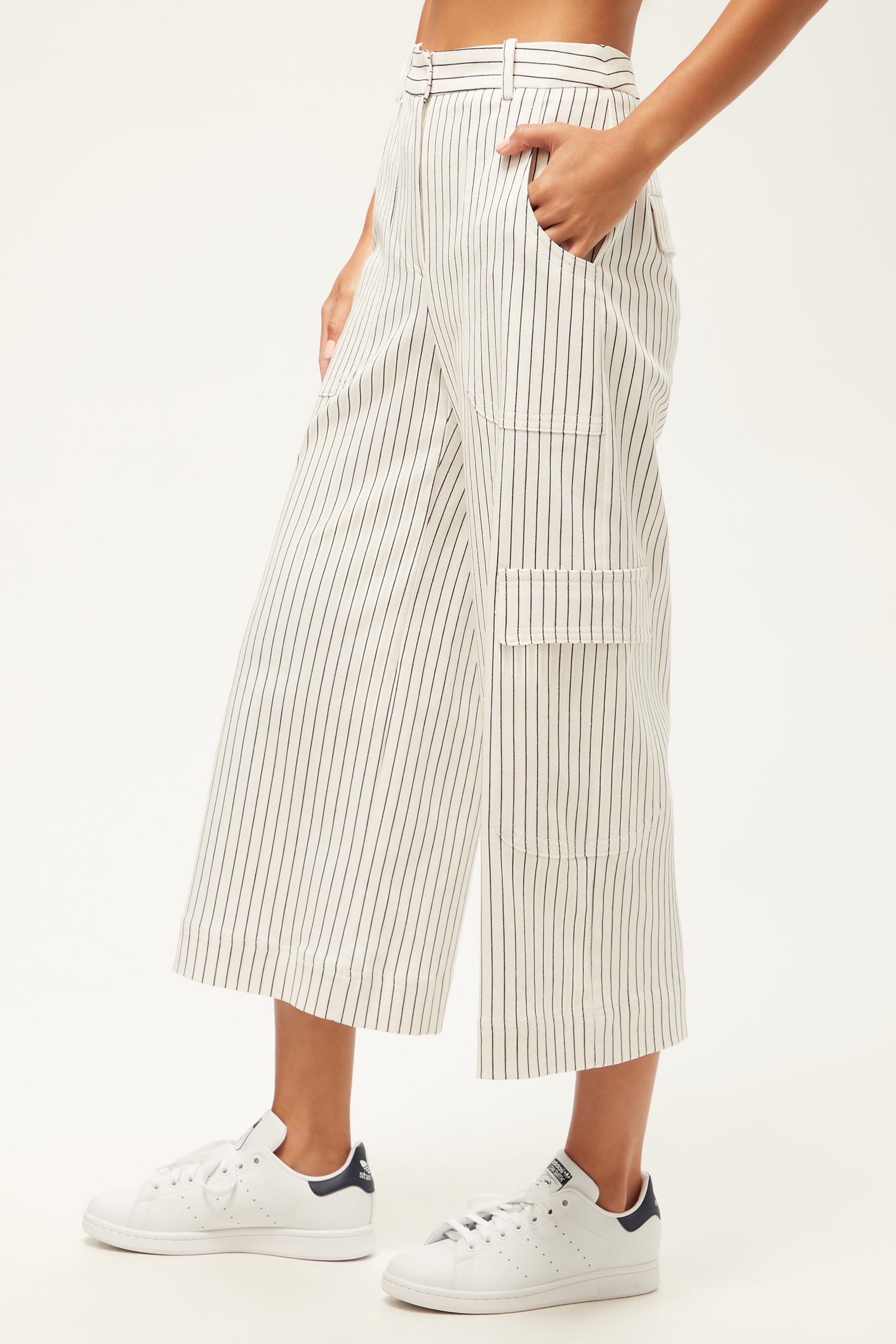A person wearing the Agassi Wide-Leg Cargo Pant - Seles Stripe, featuring high-waisted, wide-leg white pants with thin vertical black stripes and side pockets, paired with white sneakers against a plain, light background.