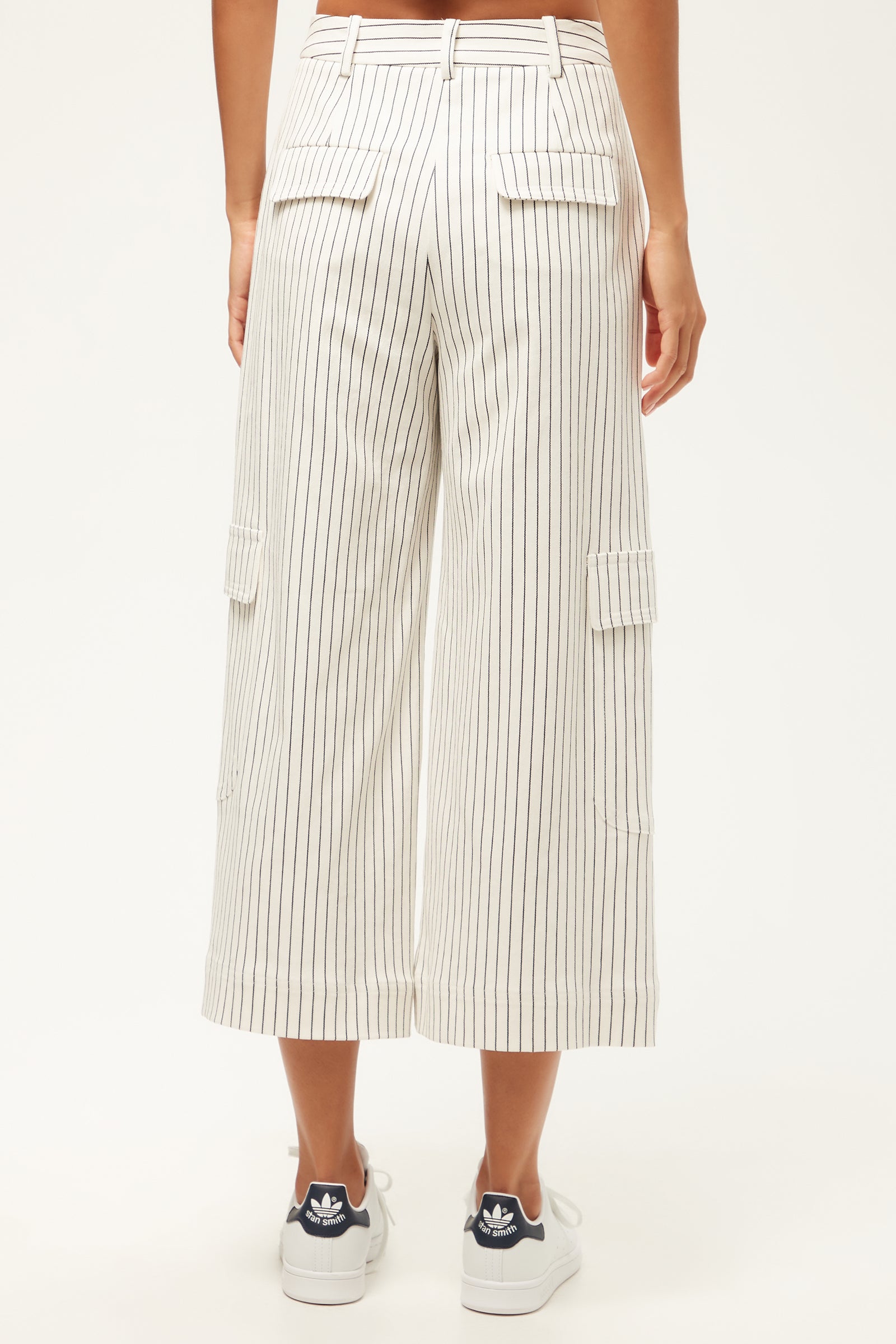 Someone stands facing away, wearing the Agassi Wide-Leg Cargo Pant in Seles Stripe—white with vertical black stripes and flap pockets—paired with white Adidas sneakers featuring navy blue accents.