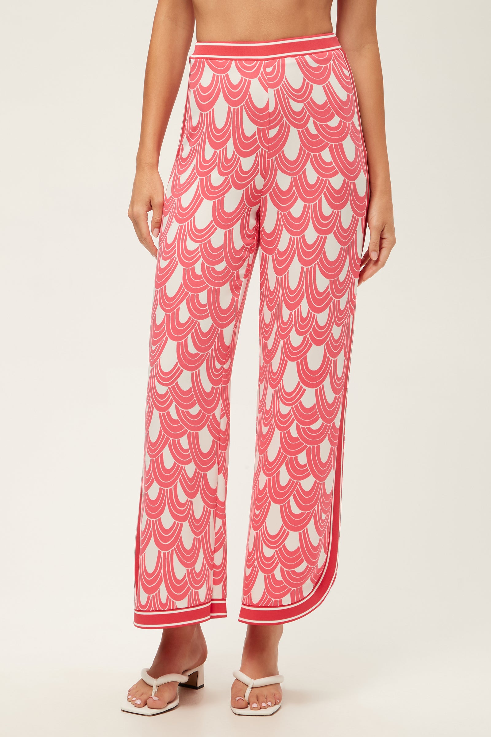 A woman showcases the Overhead Wide-Leg Pant - Ace Arches in Coral Crush/Whitewash, featuring a scalloped pattern and high-waisted, wide-leg fit. She pairs them with white heeled sandals; the image is cropped from shoulders down on a plain background.