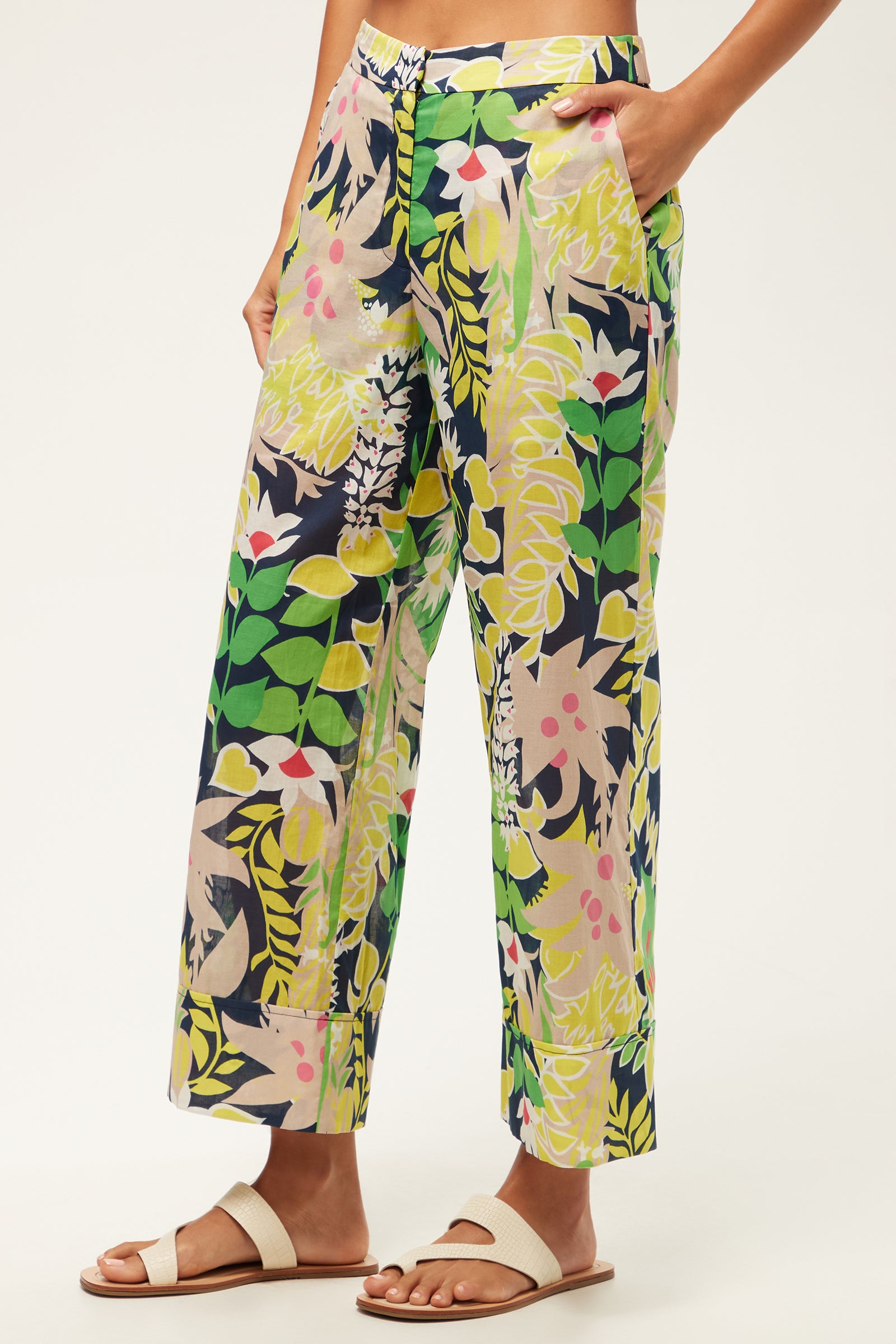 Wearing the Wilson Wide-Leg Pant - PS Eternal featuring a vibrant green, yellow, navy, and pink floral and leaf print, paired with white sandals. One hand is in the pocket against a plain light background.