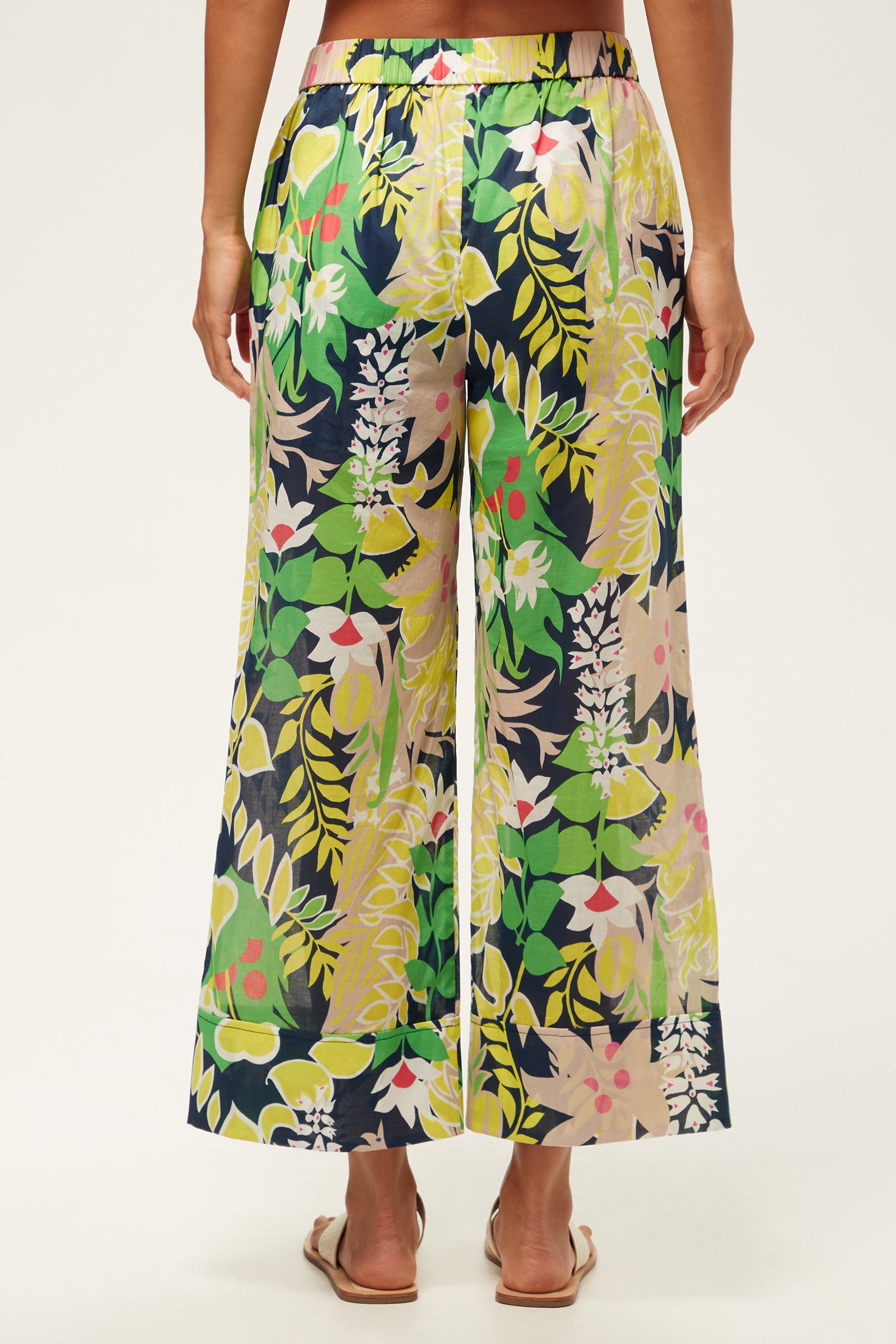 A person is shown from the waist down, wearing the Wilson Wide-Leg Pant - PS Eternal with a vibrant floral and leaf print in green, yellow, pink, and navy, paired with flat sandals against a white background.