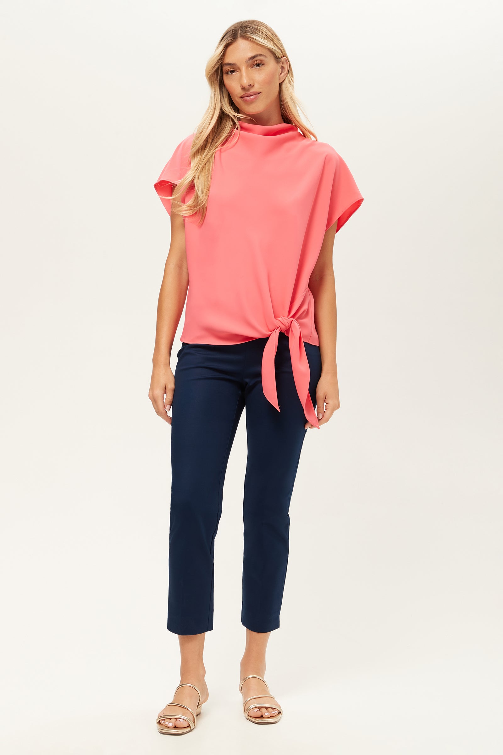 A woman with long blonde hair and a relaxed expression wears a bright pink short-sleeve blouse, beige sandals, and the Tie Breaker Slim Crop Pant - Solid Suiting in navy blue against a plain background.
