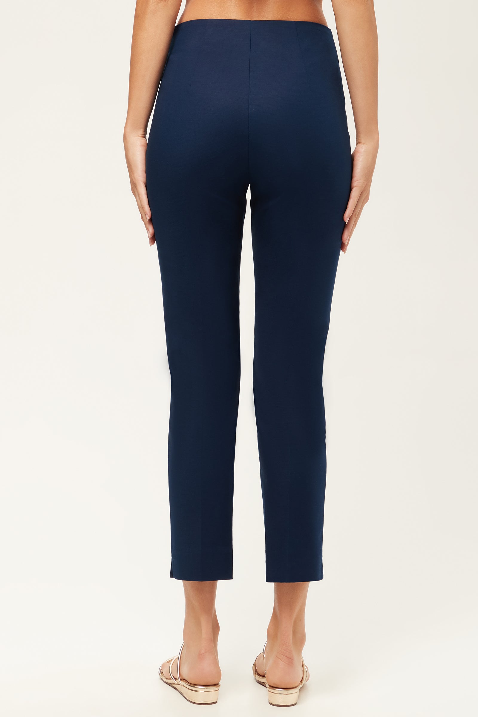 A person stands facing away, wearing the Tie Breaker Slim Crop Pant - Solid Suiting in navy blue and beige sandals, with hands at their sides against a plain light background.