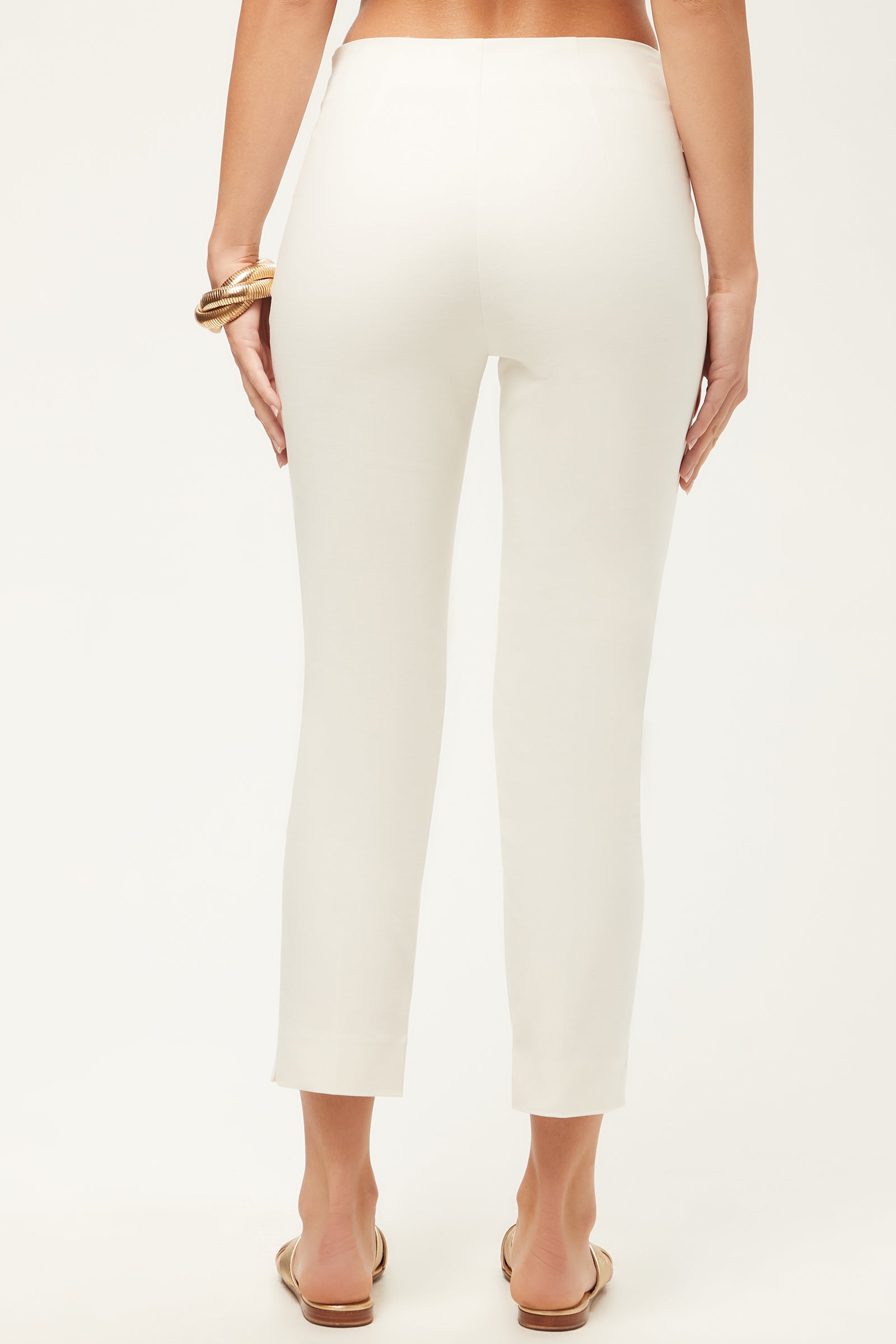 A person is shown from the back wearing white Tie Breaker Slim Crop Pant - Solid Suiting and beige slide sandals, accessorized with a gold bangle, standing against a plain light background.