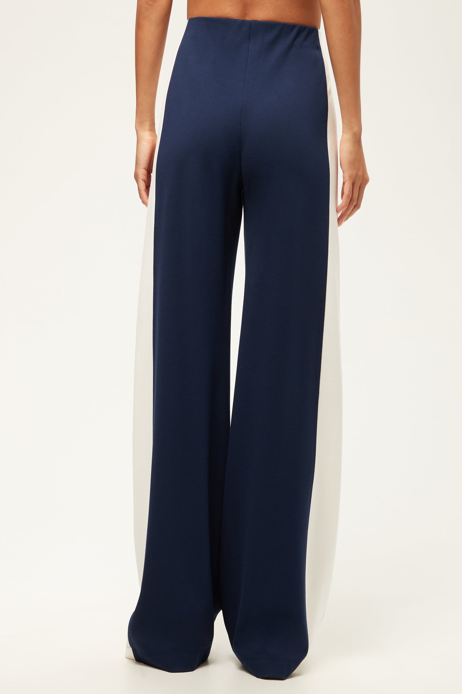 A person stands with their back to the camera, wearing the Record Wide-Leg Pant - Cozy Ponte in navy, featuring high-waisted, wide-leg design and white side panels against a plain light background.