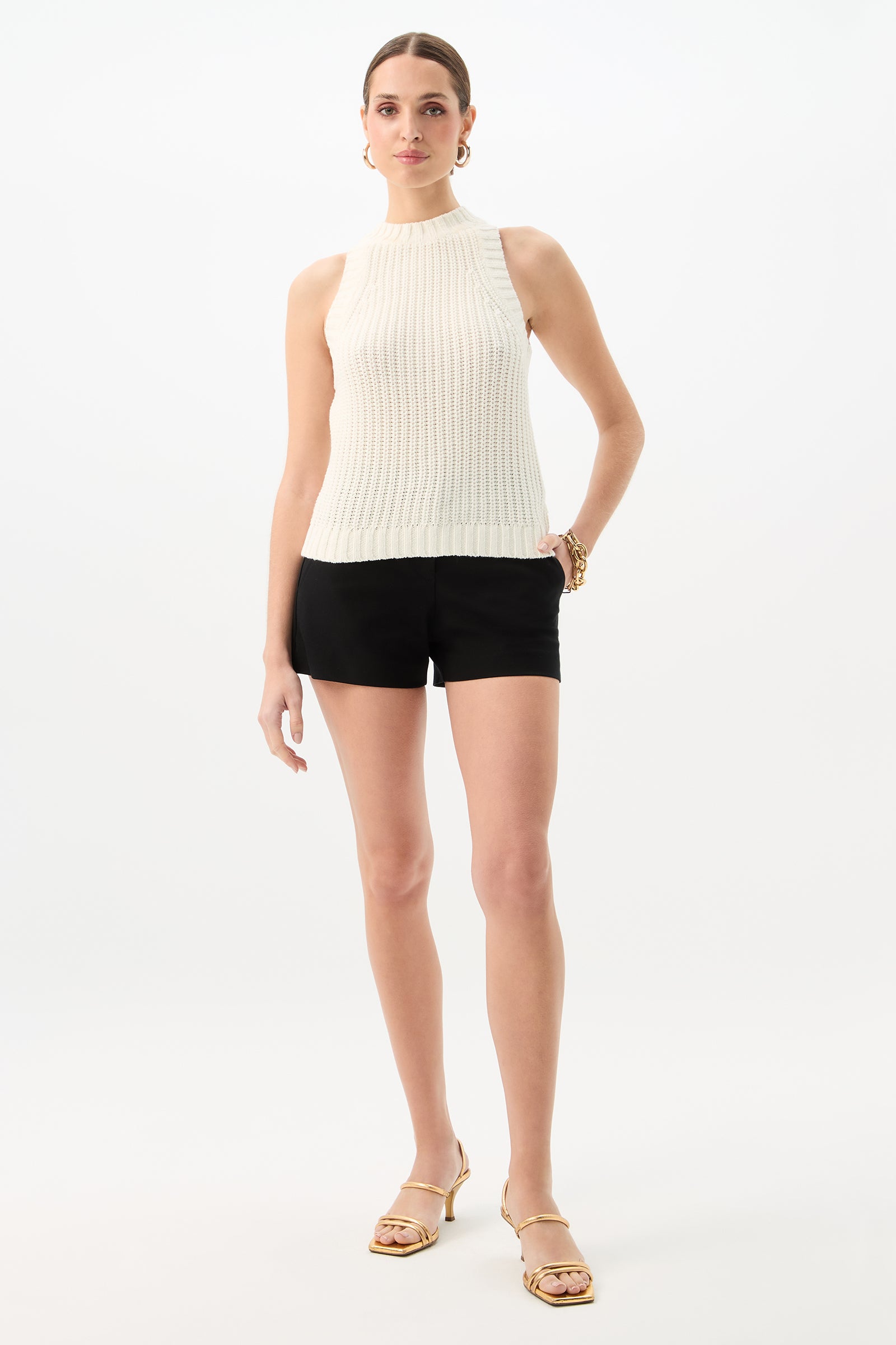 A woman stands against a white background wearing the Paley Sleeveless Rib Knit Tank Top, black shorts, gold hoop earrings, a gold bracelet, and gold sandals. Her hair is pulled back and one hand rests on her hip.
