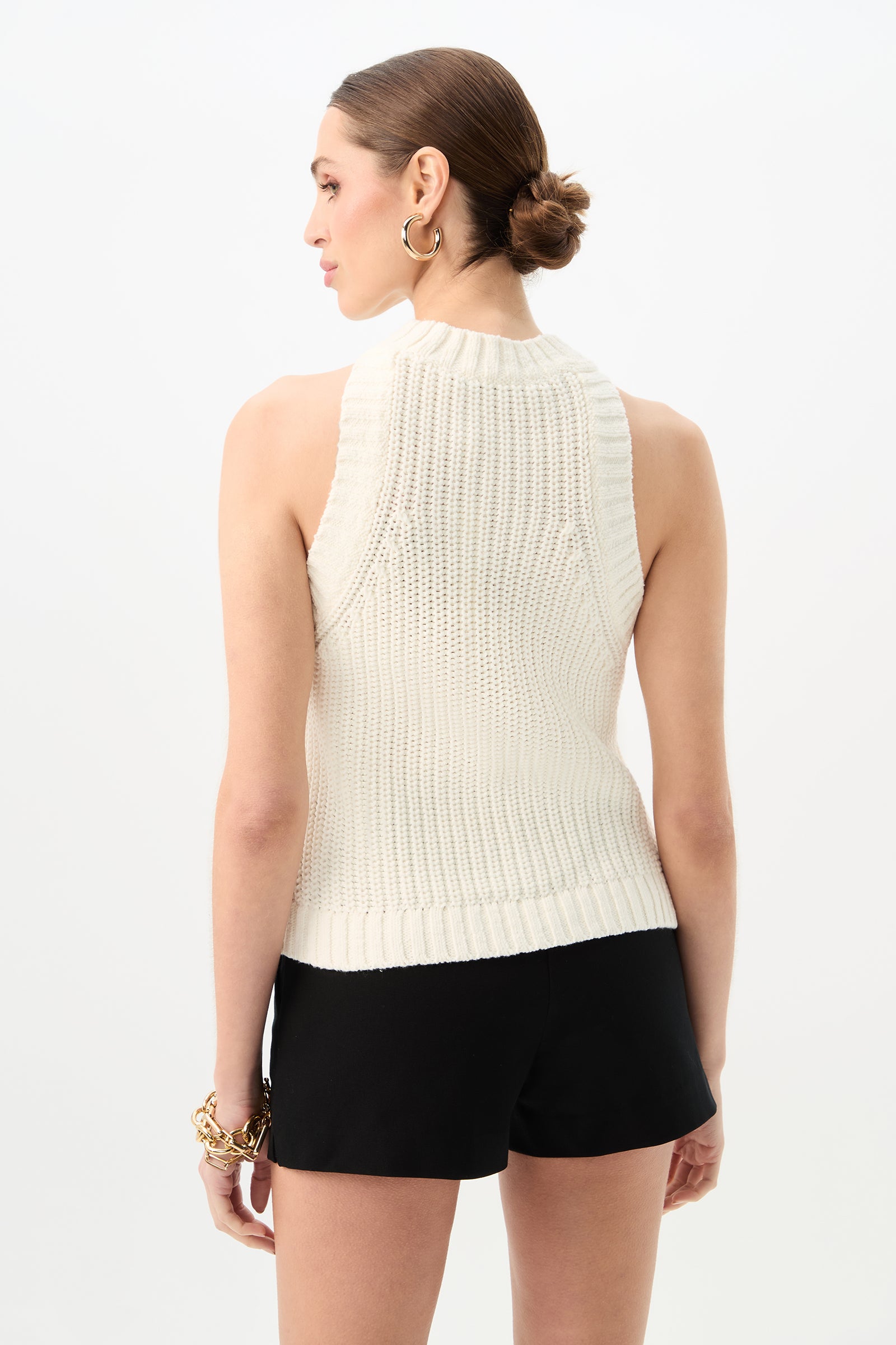 A woman with brown hair in a low bun wears the Paley Sleeveless Rib Knit Tank Top in white and black shorts, with her back to the camera. She accessorizes with gold hoop earrings and a chunky gold bracelet.