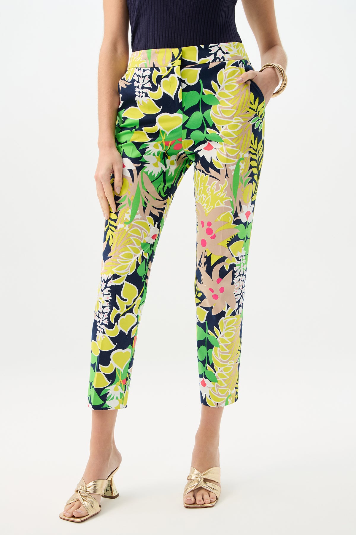 Wearing the Aubree 2 Pant - PS Eternal, a person poses with hands in pockets. The high-waisted, ankle-length pants feature a multicolored floral print, paired with a navy sleeveless top and gold open-toe heeled sandals.