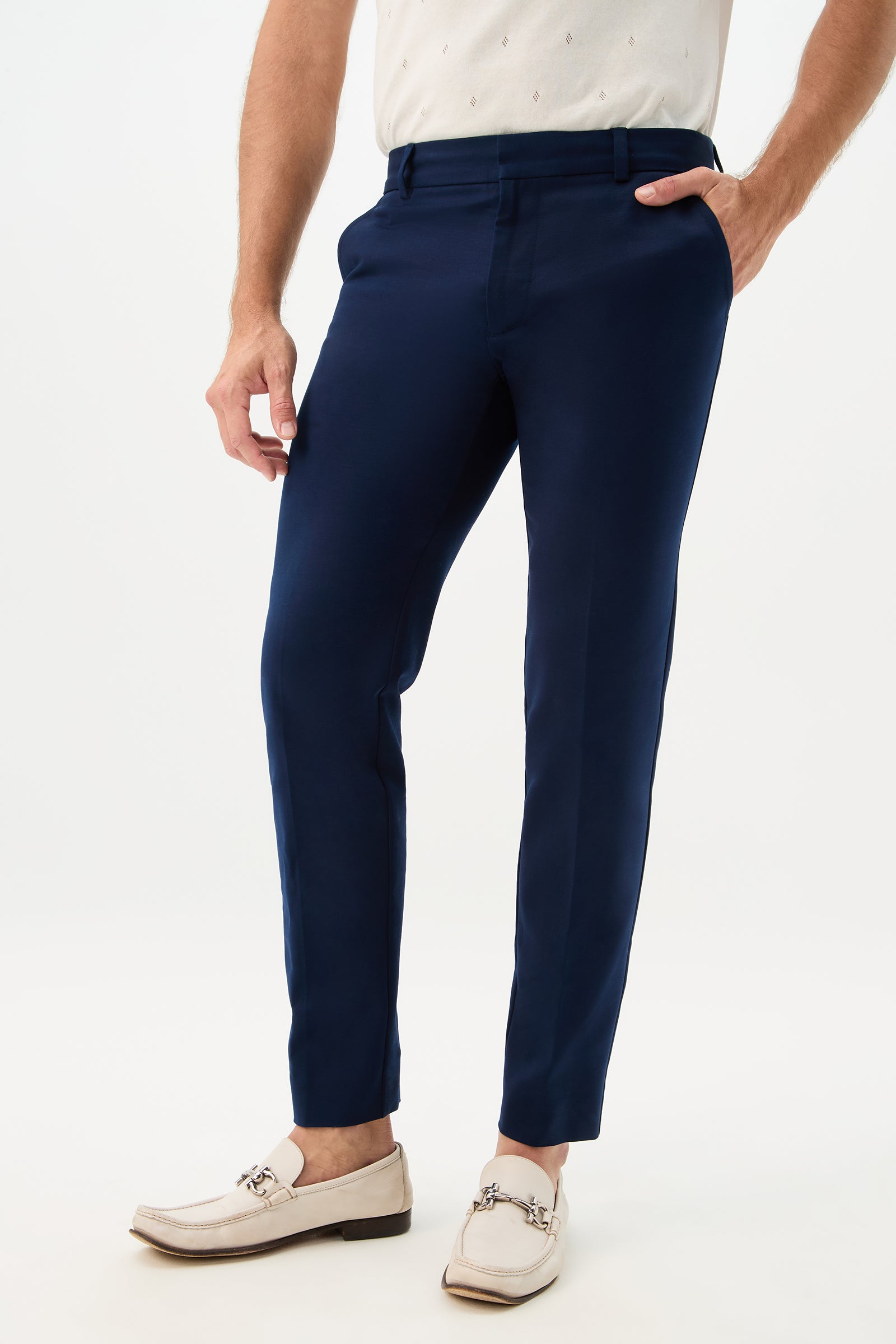 A person wears the Clyde Slim Trouser - Solid Suiting in navy, paired with a white patterned tucked shirt and cream loafers with metal accents, standing against a plain white background, one hand in their pocket.