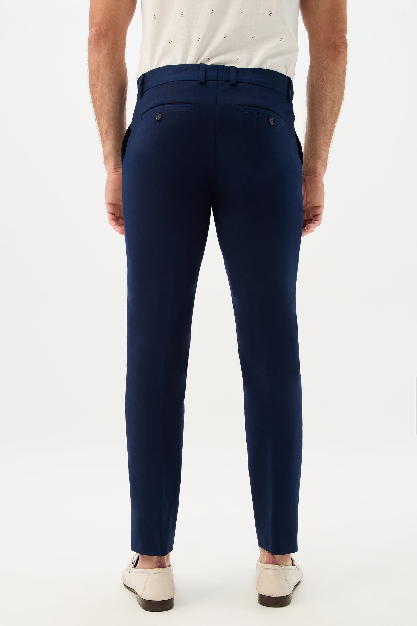 A man shown from the back against a plain white background wears the Clyde Slim Trouser - Solid Suiting in navy blue, paired with a white short-sleeve shirt and light beige loafers.