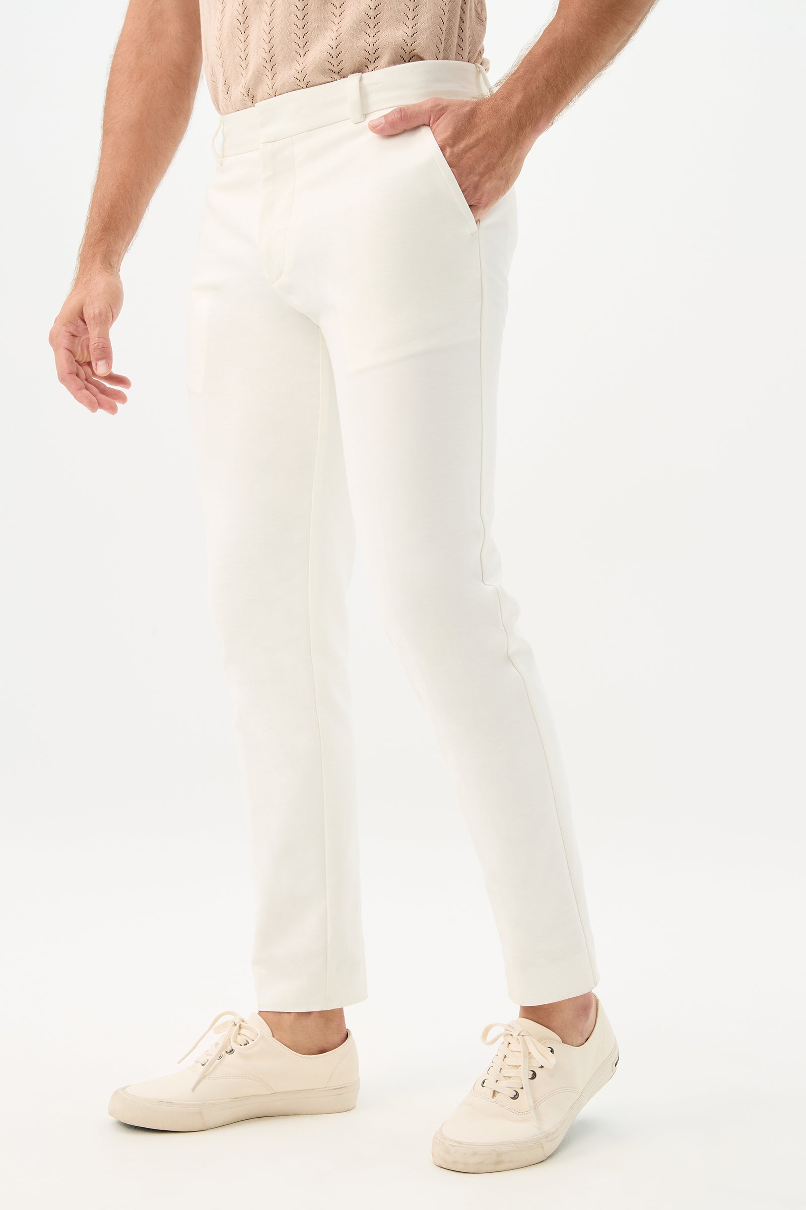 A person stands with one hand in their pocket, wearing the Clyde Slim Trouser - Solid Suiting in white, paired with a light beige knit shirt and off-white sneakers against a plain white background.