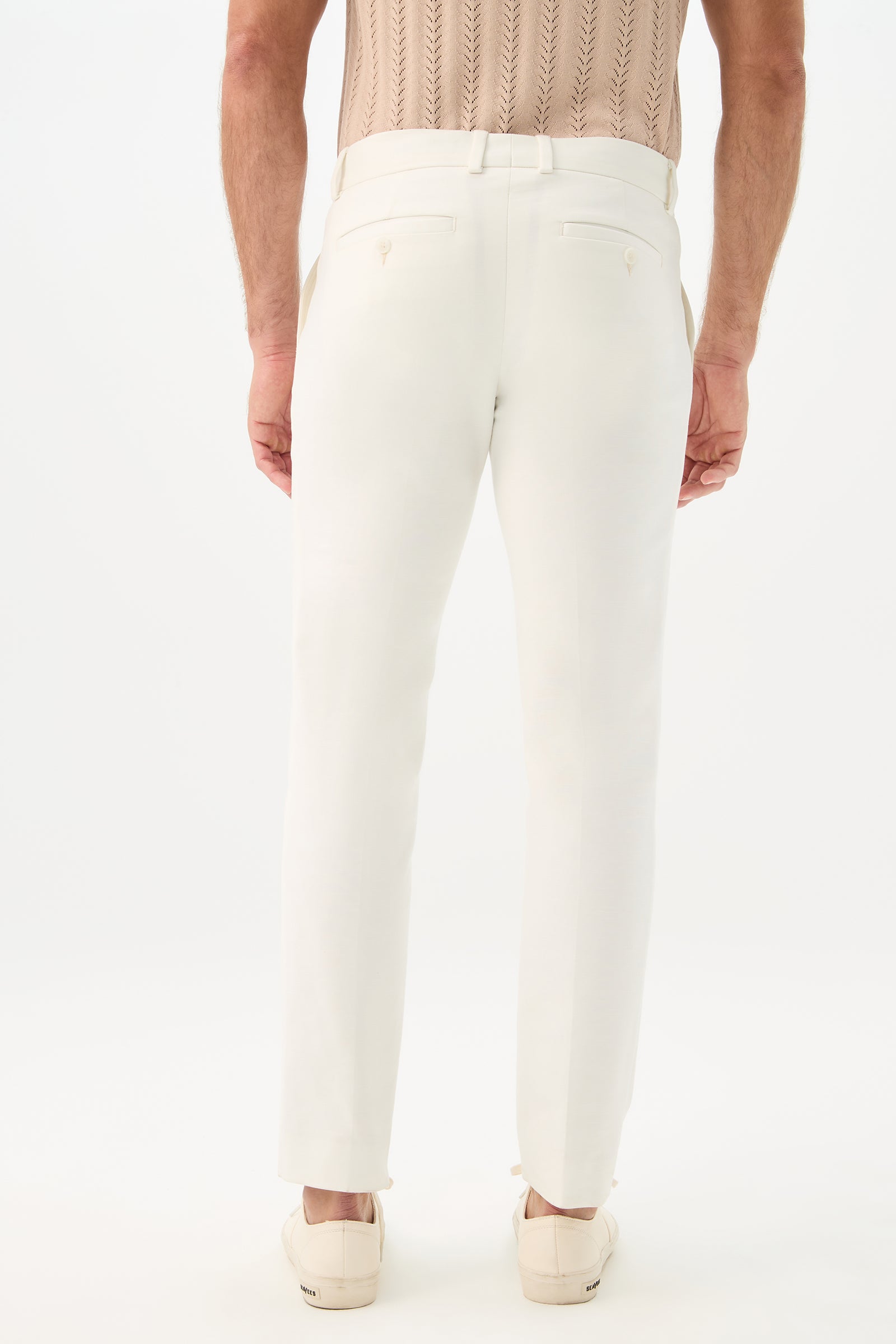 A man faces away from the camera, wearing Clyde Slim Trouser - Solid Suiting in white, a beige short-sleeve knit shirt, and beige slip-on shoes against a plain white background.