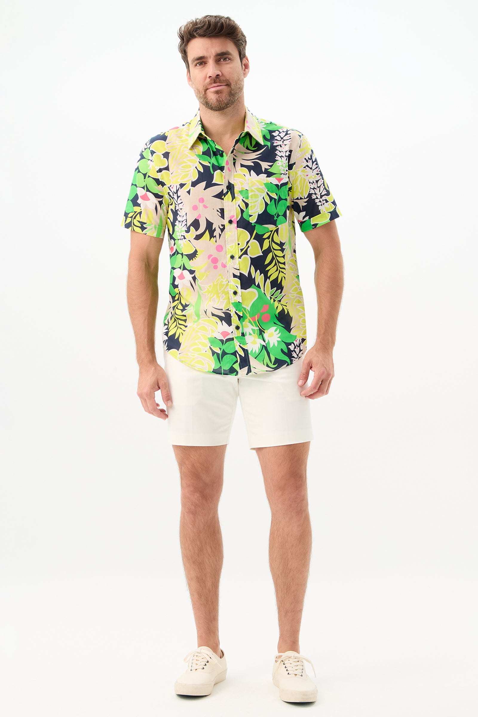 A man with short hair and a beard stands against a white background wearing the Lawrence Short - Solid Suiting, paired with a floral short-sleeve shirt and white sneakers. He appears relaxed, with his hands by his sides.