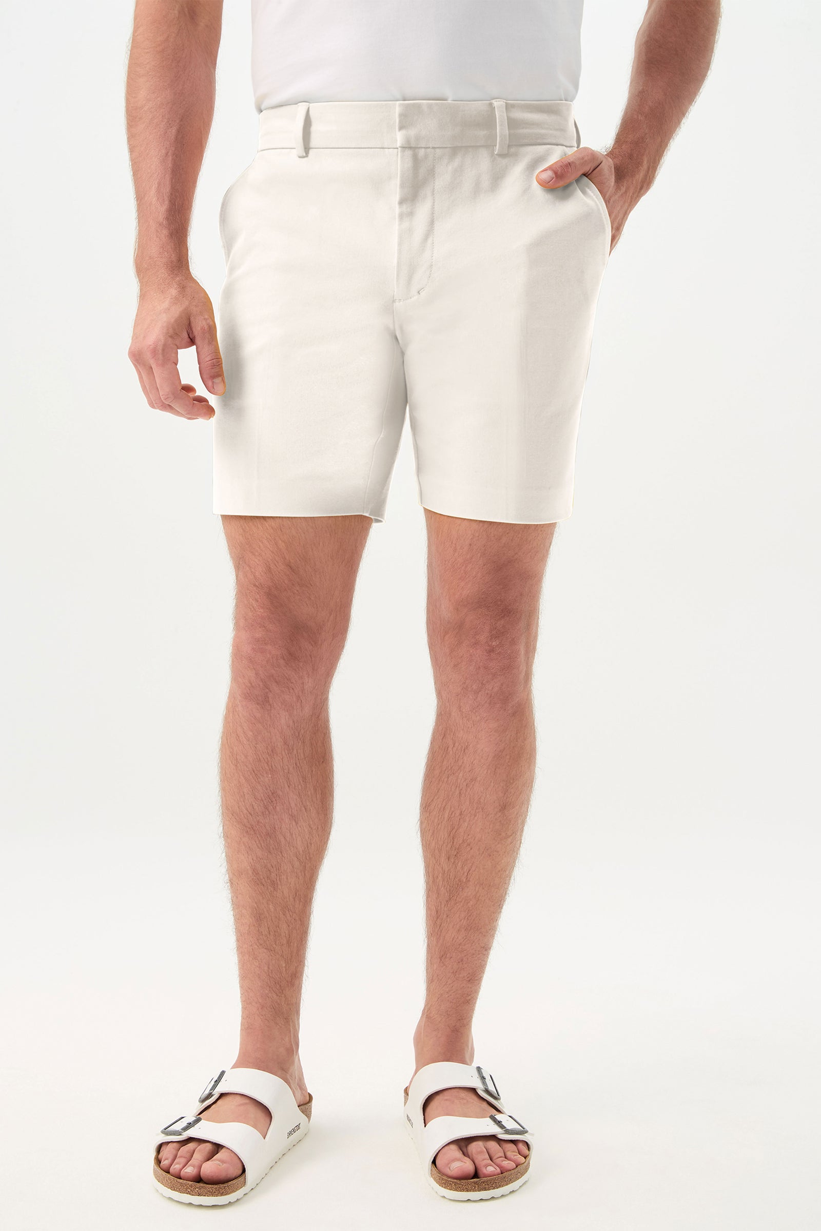 A person wearing the Lawrence Short - Solid Suiting in Whitewash, paired with a white t-shirt and two-strap white sandals, stands with one hand in their pocket against a plain light background.