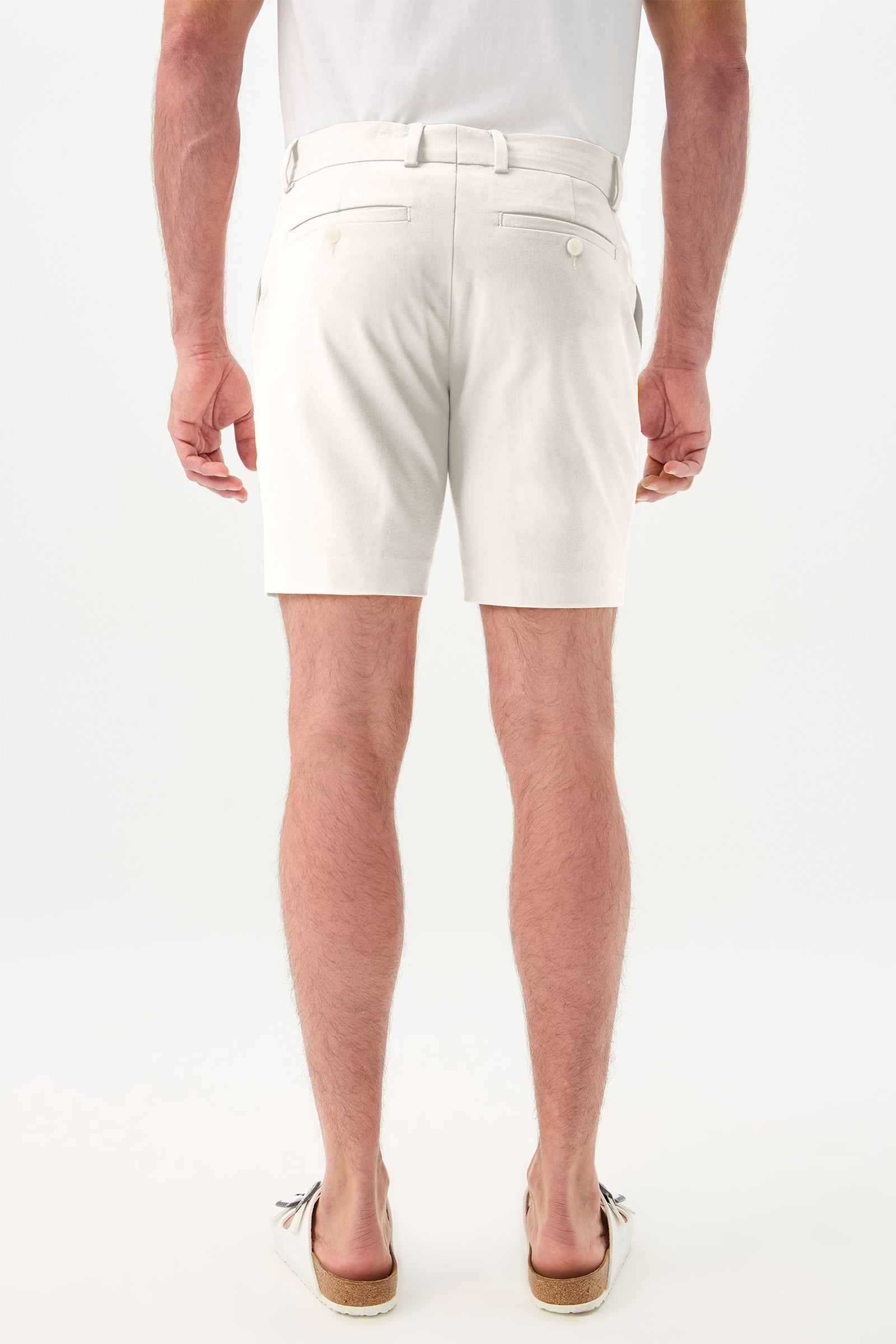 A man stands with his back to the camera, wearing a white t-shirt, Lawrence Short - Solid Suiting with back pockets, and beige tassel loafers. The background is plain white.