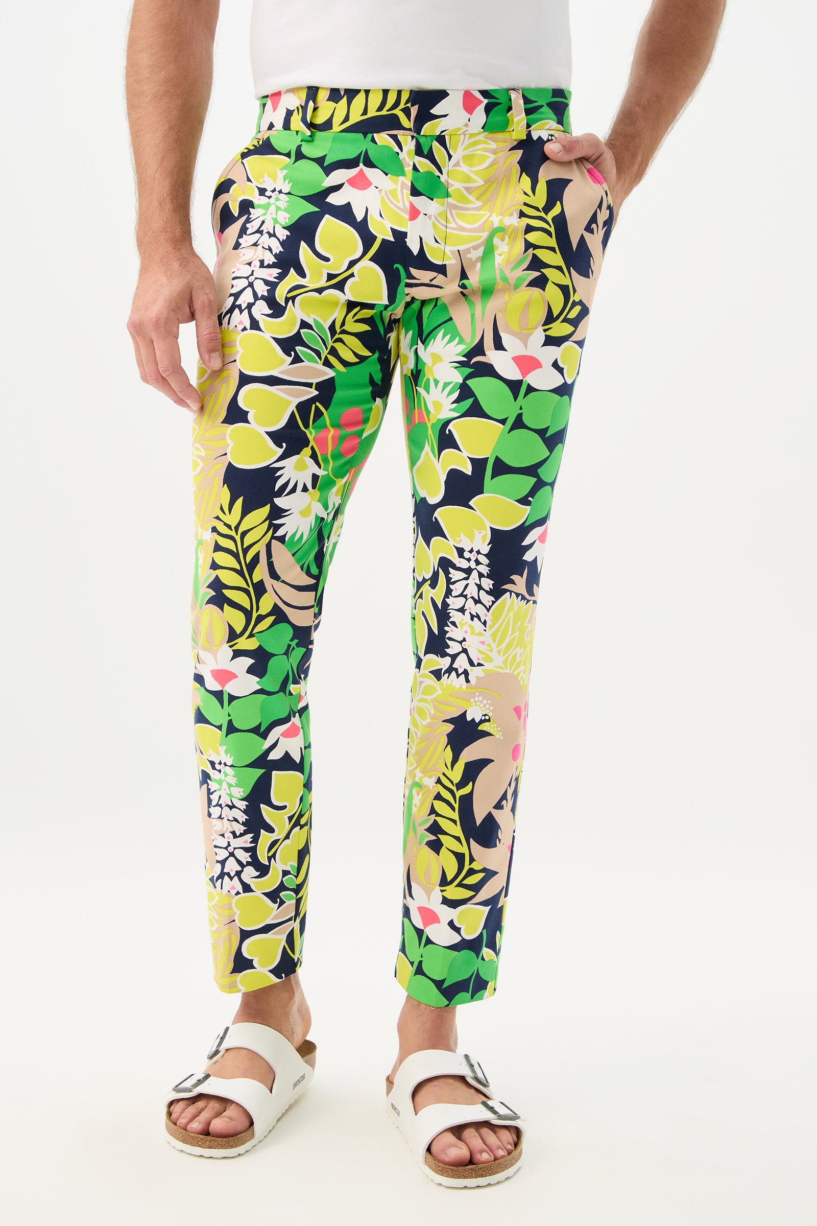 A person wearing the Clyde Slim Trouser - PS Eternal, featuring green, yellow, pink, white, and navy tropical floral patterns, paired with a white shirt and white sandals, stands against a plain white background.