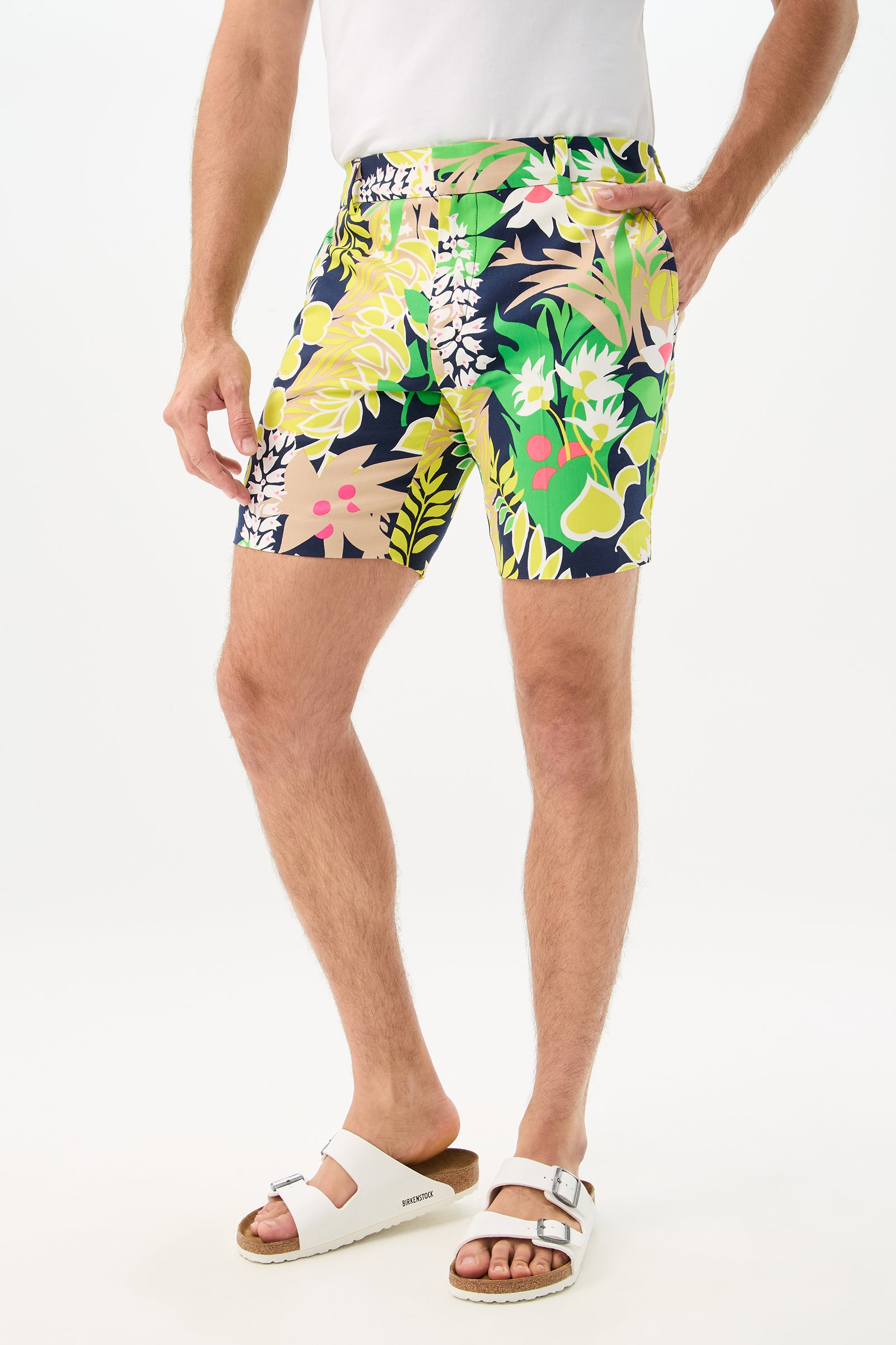 A person models the Lawrence Short - PS Eternal, featuring colorful tropical patterns, paired with a white shirt and white sandals, standing with one hand in their pocket against a plain white background.