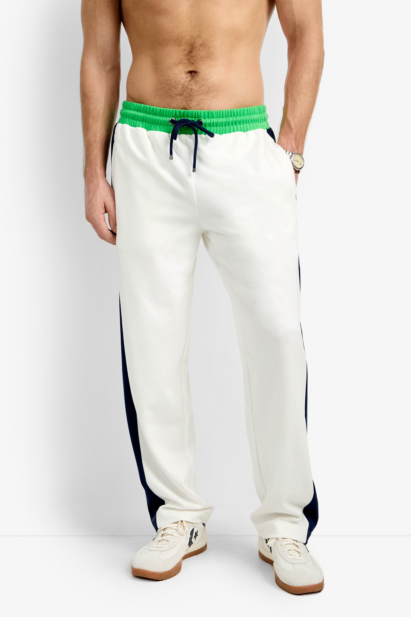 A shirtless man wearing the Tennis Track Pant - Cozy Ponte in white with a green waistband, black side stripes, and white sneakers stands against a plain background.
