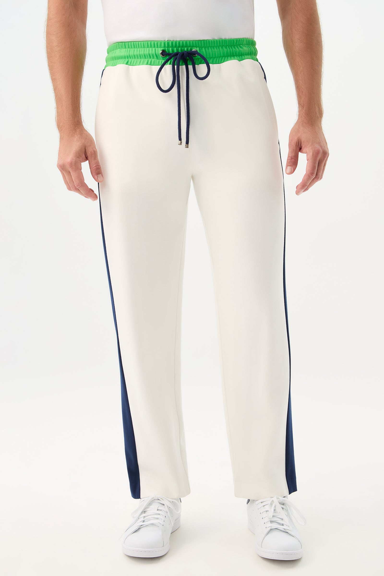 A person wears the Tennis Track Pant - Cozy Ponte in Multi, featuring a green waistband, navy blue drawstring and side stripes, paired with white sneakers, standing against a plain white background.
