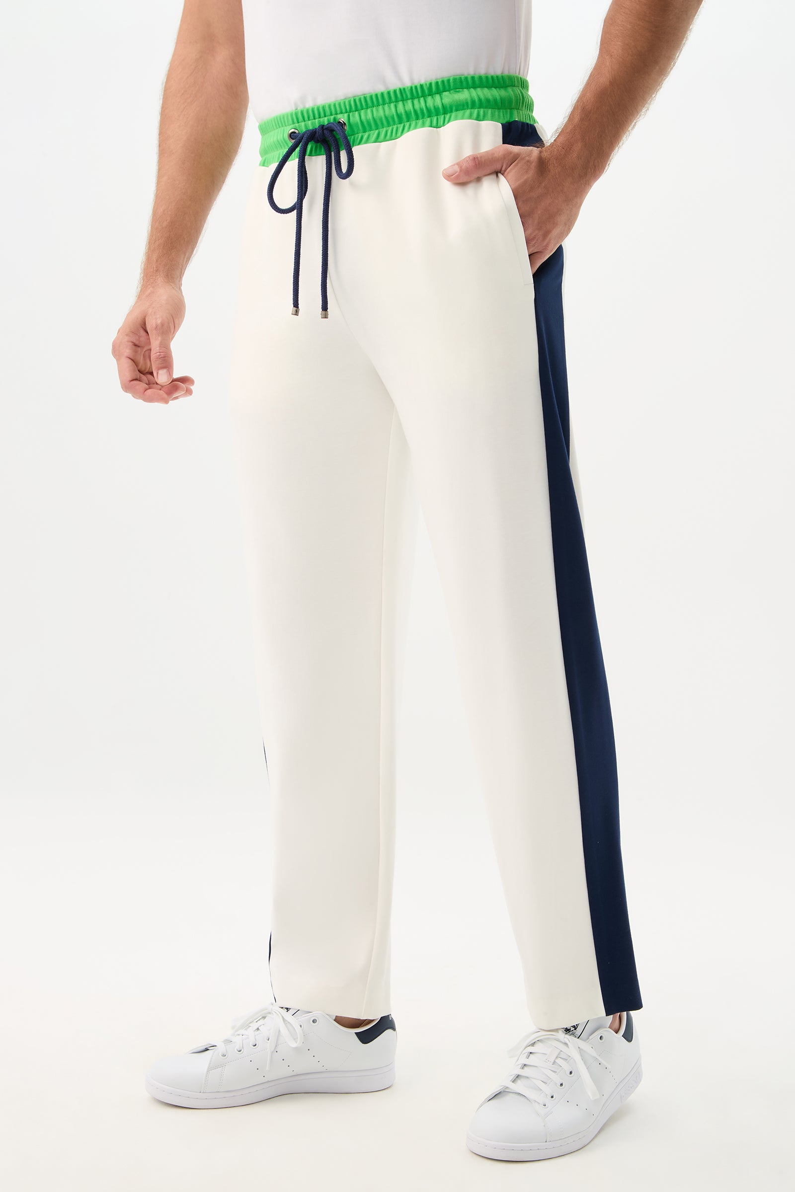 A person models the Tennis Track Pant - Cozy Ponte, featuring white fabric, a green waistband, navy side stripes, black drawstring, and white sneakers against a plain white background.